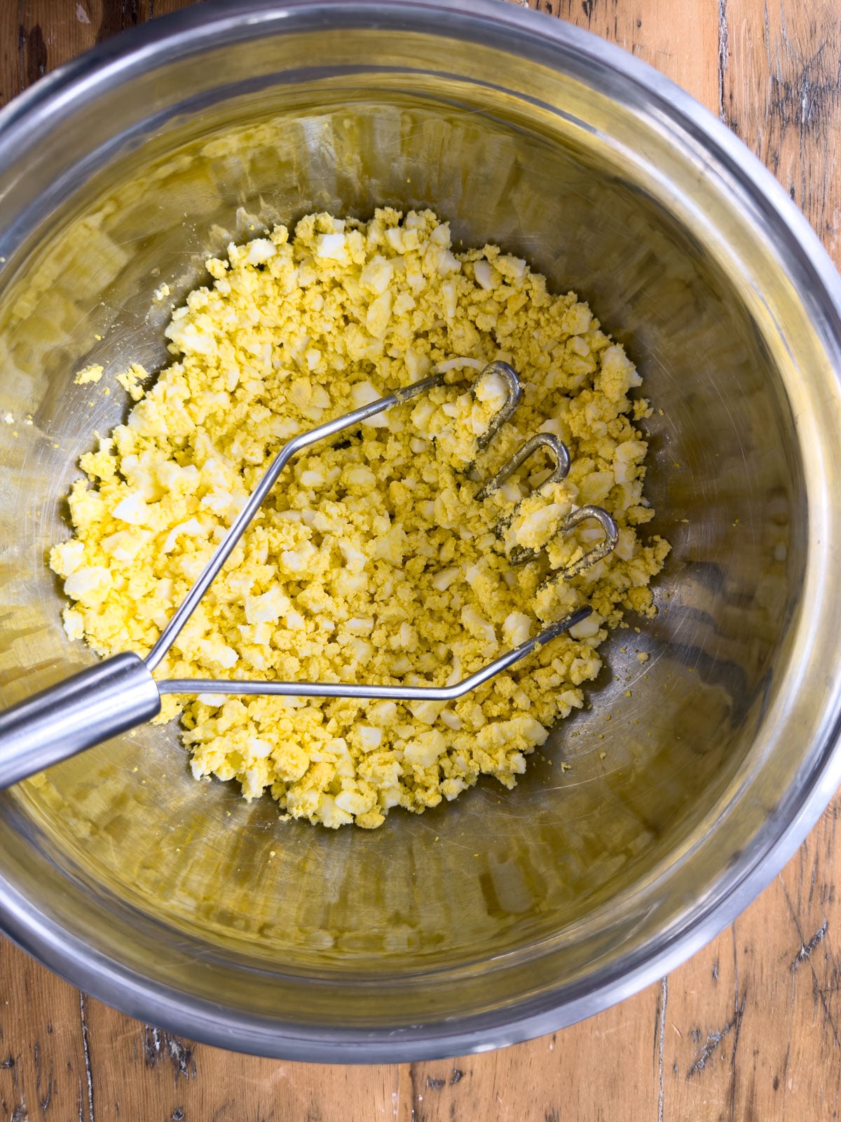 Hard boiled eggs mashed with a potato masher in a stainless steel mixing bowl.