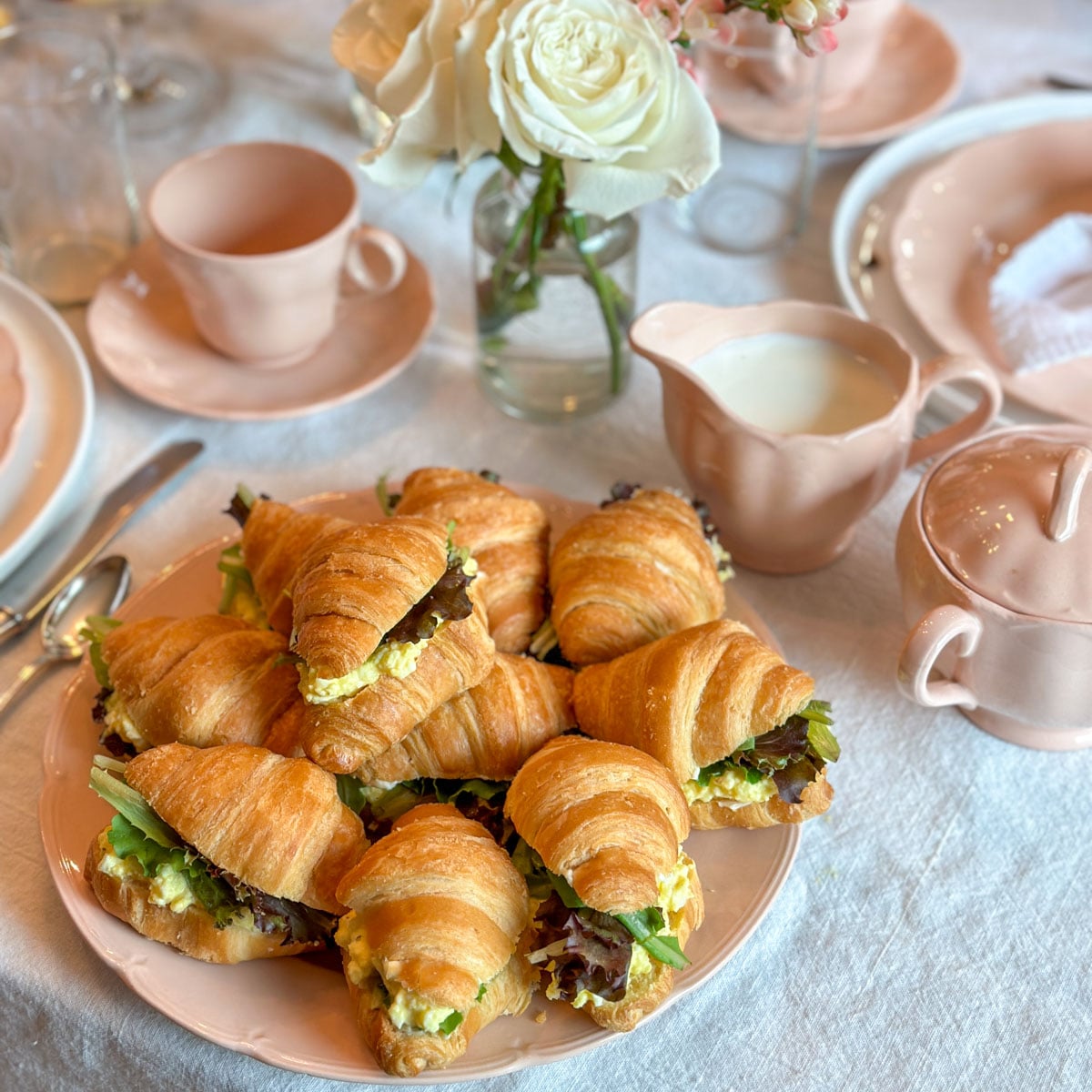 Mini egg salad croissant sandwiches with mixed greens piled on a pink serving plate surrounded by a pink tea service and white roses.