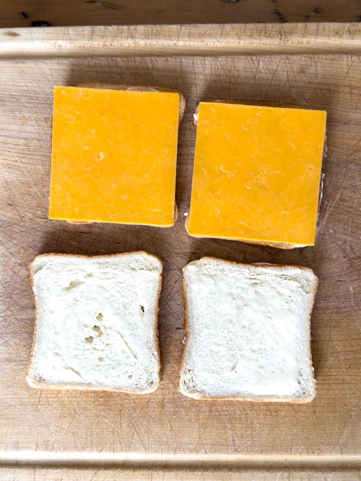 Two slices of buttered white bread and two slices topped with cheddar cheese on a wooden cutting board.