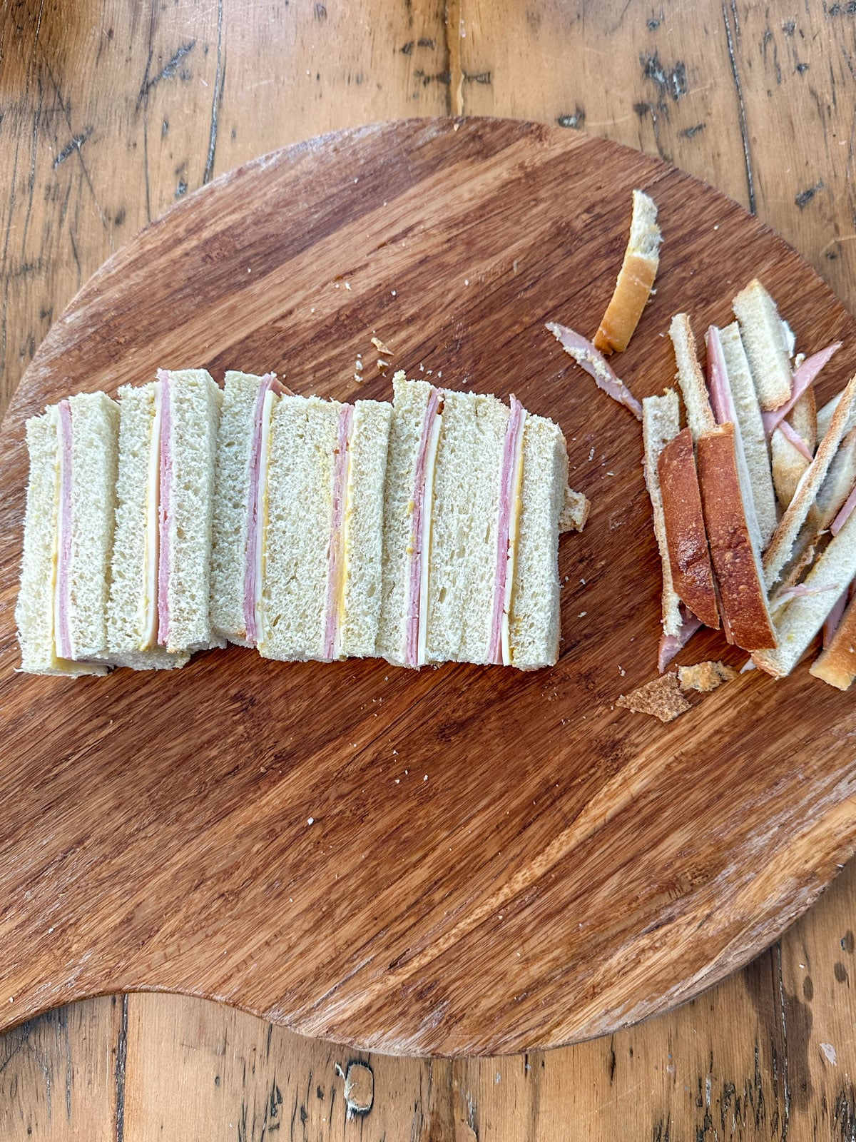 Honey ham and Swiss finger sandwiches cut into rectangular fingers on a round wooden board with the crusts set to the side.