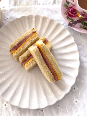 Three ham and cheddar tea sandwich fingers on a white scalloped plate with a rose teacup and stacked vintage plates in the background.
