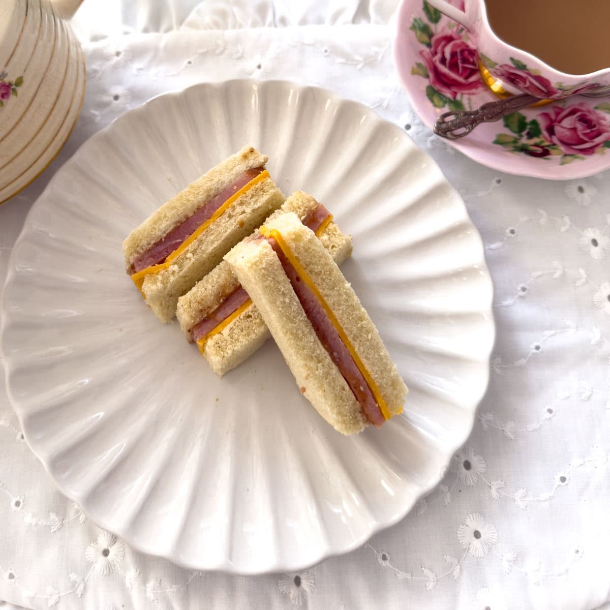 Three ham and cheddar tea sandwich fingers on a white scalloped plate with a rose teacup and stacked vintage plates in the background.