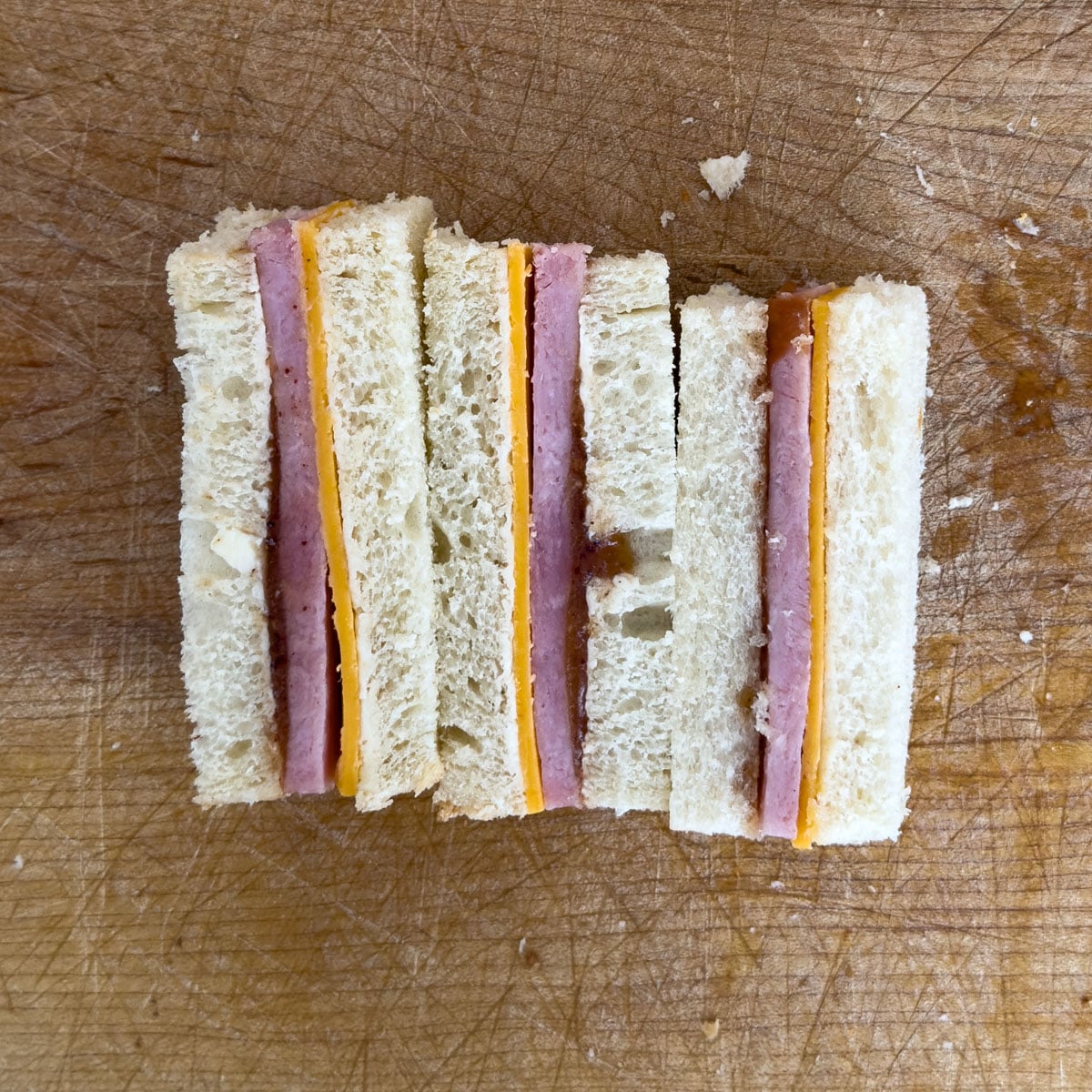 Ham and cheddar tea sandwich fingers cut and arranged on a wooden cutting board with the crusts removed.