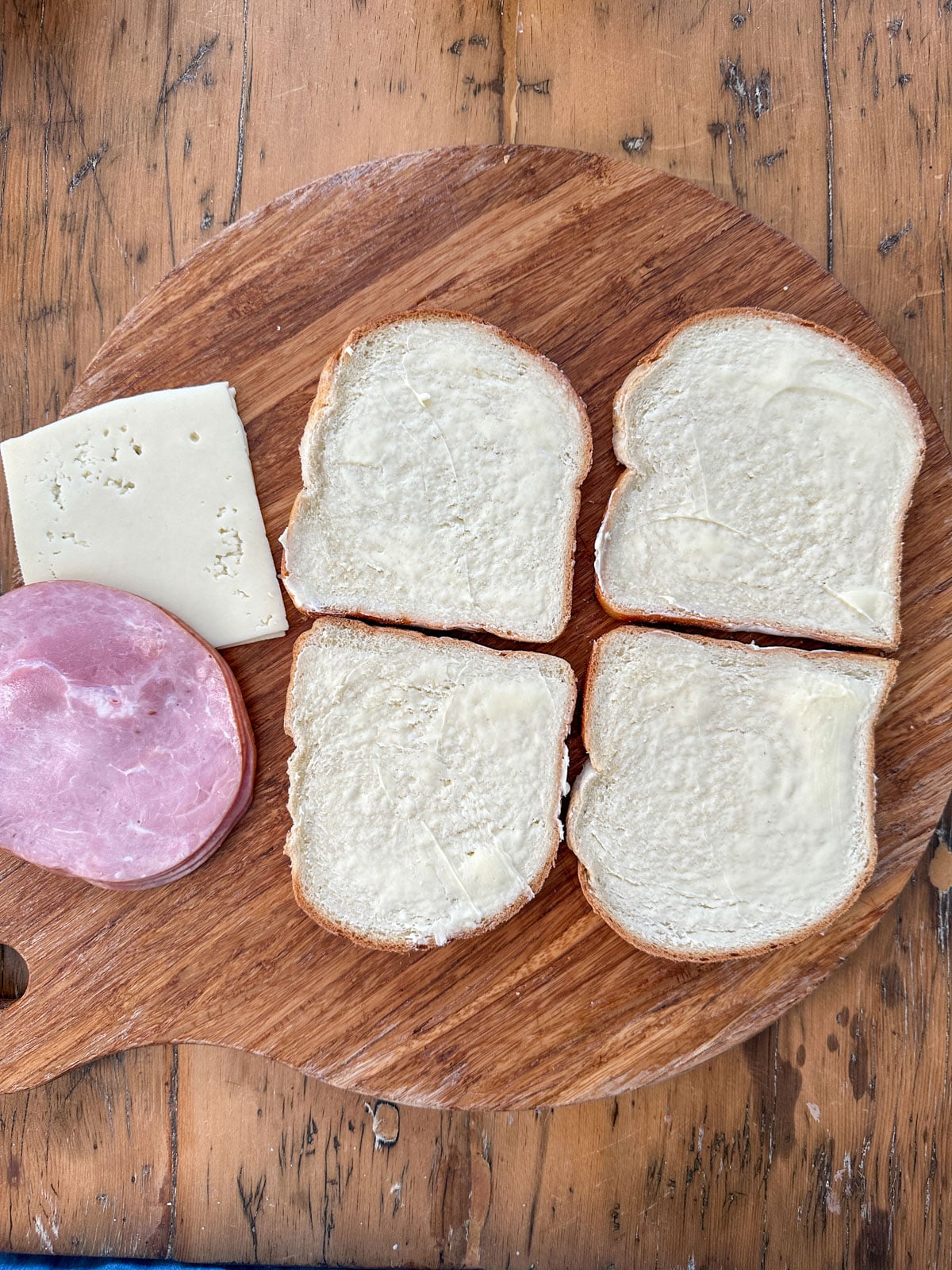 Four buttered slices of white sandwich bread on a round wooden board with sliced honey ham and Swiss cheese set to the side.