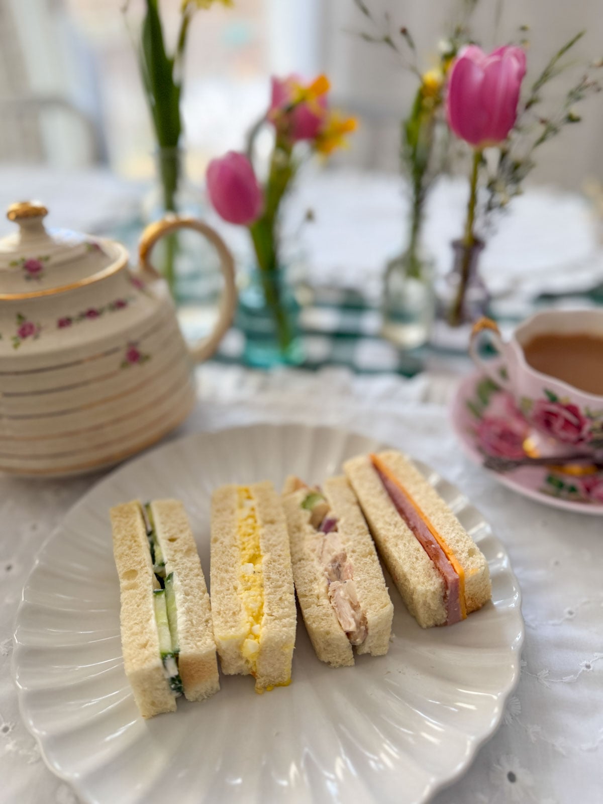 Cucumber, egg salad, chicken salad and ham and cheddar tea sandwich fingers on a white scalloped plate with a rose teapot, pink rose teacup, tulips and spring flowers on a green gingham tablecloth.