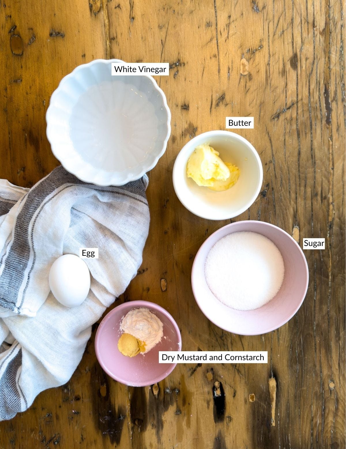Homemade salad dressing ingredients including white vinegar, butter, egg, sugar, dry mustard, and cornstarch labeled on a rustic wood surface.