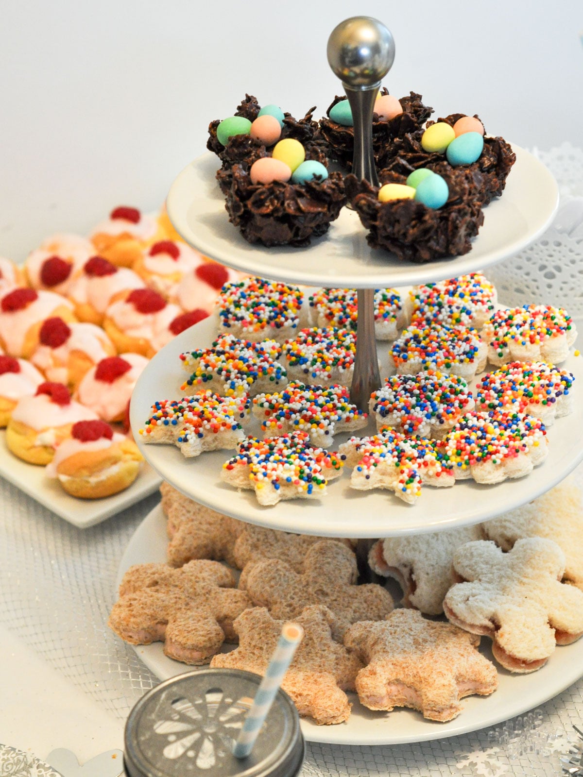 A tiered stand displaying fairy bread cut into star shapes and chocolate bird's nest treats with candy eggs, beside a plate of whole wheat teddy bear shaped sandwiches and pink frosted cream puffs topped with raspberries.