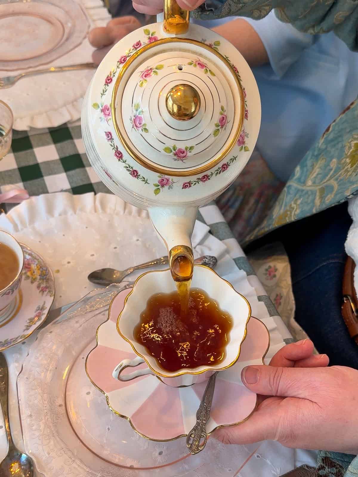 Vintage rose-patterned teapot pouring amber tea into a gold-rimmed teacup at a Little Women vintage tea party.