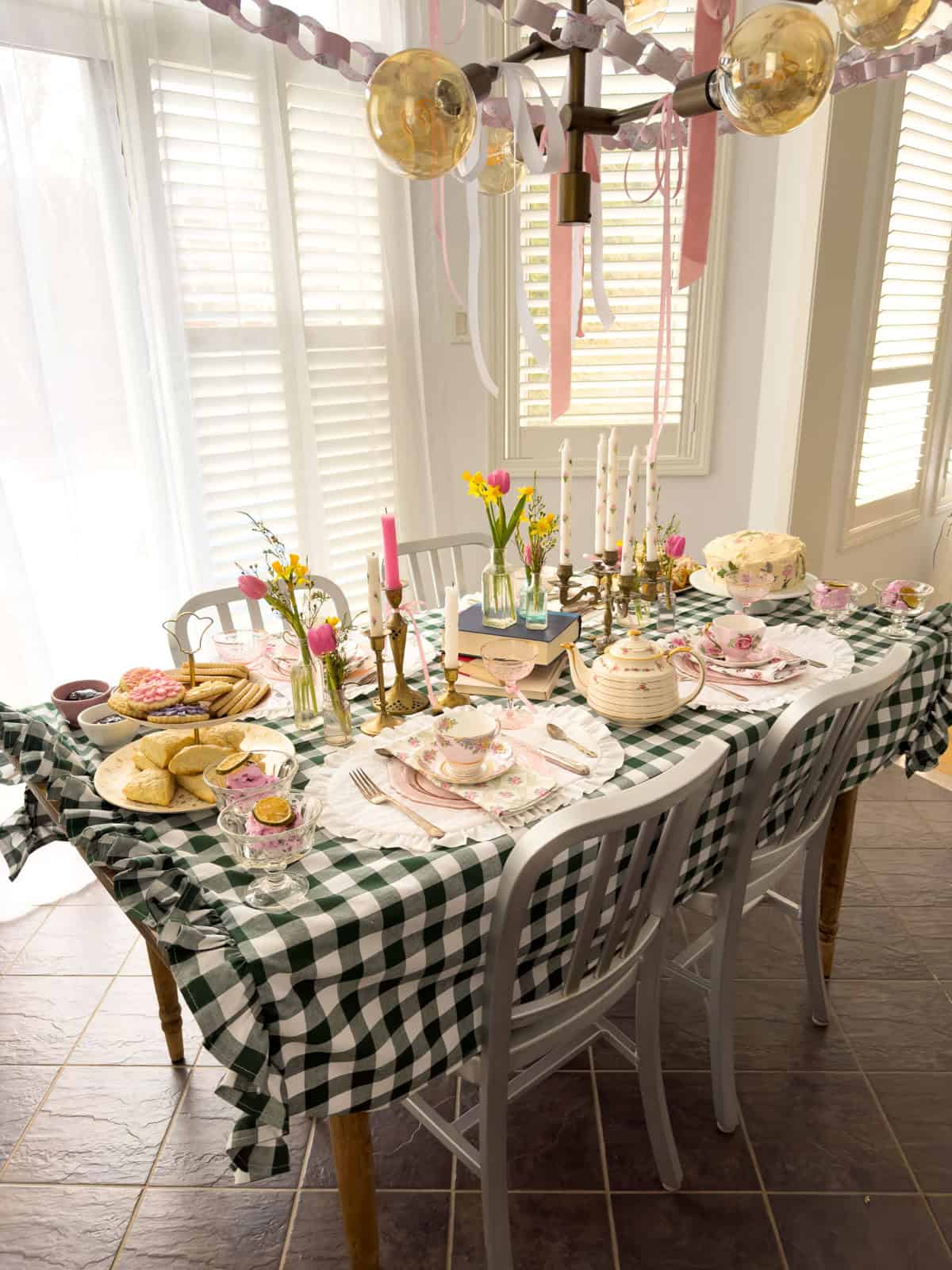 Full table spread at a Little Women vintage tea party with scones, sugar cookies, pressed flower cake, vintage teapots, brass candelabra, spring flowers and paper chains overhead on a green gingham tablecloth