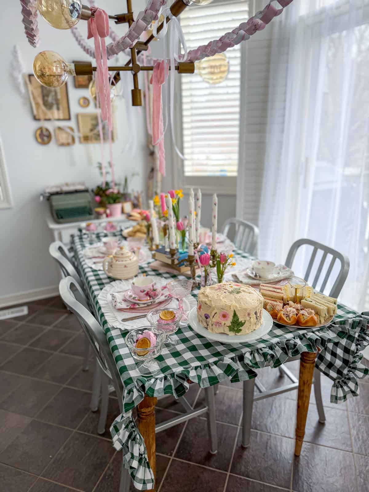 Little Women vintage tea party menu spread with pressed flower cake, chicken salad finger sandwiches and ice cream cups on a green gingham table with hand-painted candles and spring flowers.