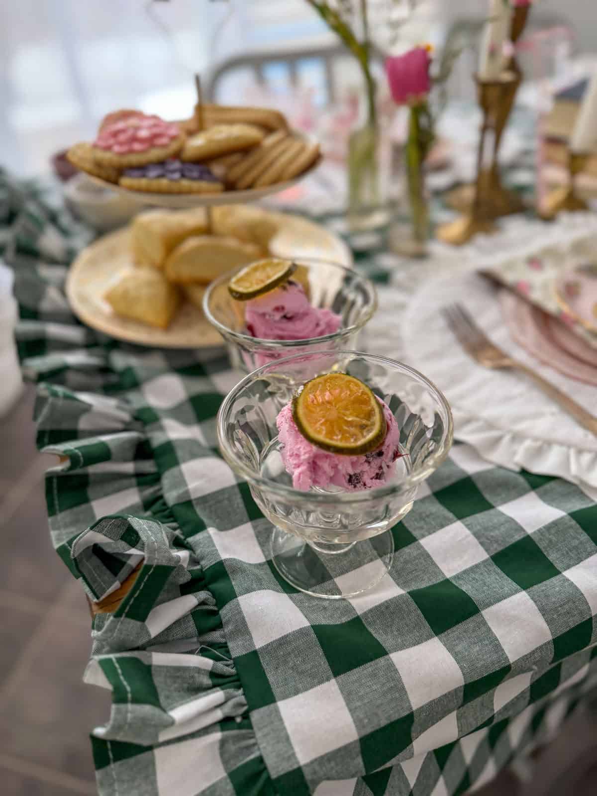 Pink strawberry ice cream topped with a candied lime slice in a vintage glass dish at a Little Women vintage tea party with scones and sugar cookies behind.