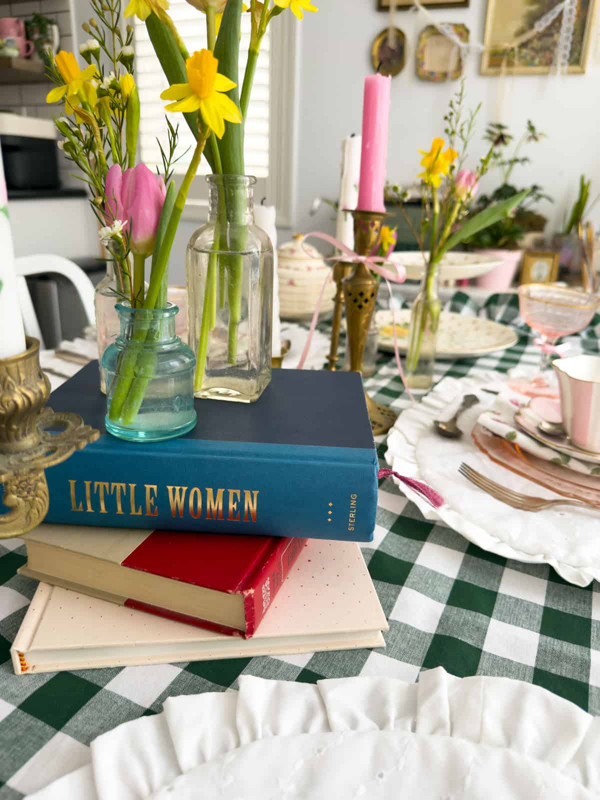 Blue hardcover Little Women book stacked with vintage books on a green gingham tablecloth surrounded by daffodils and tulips.