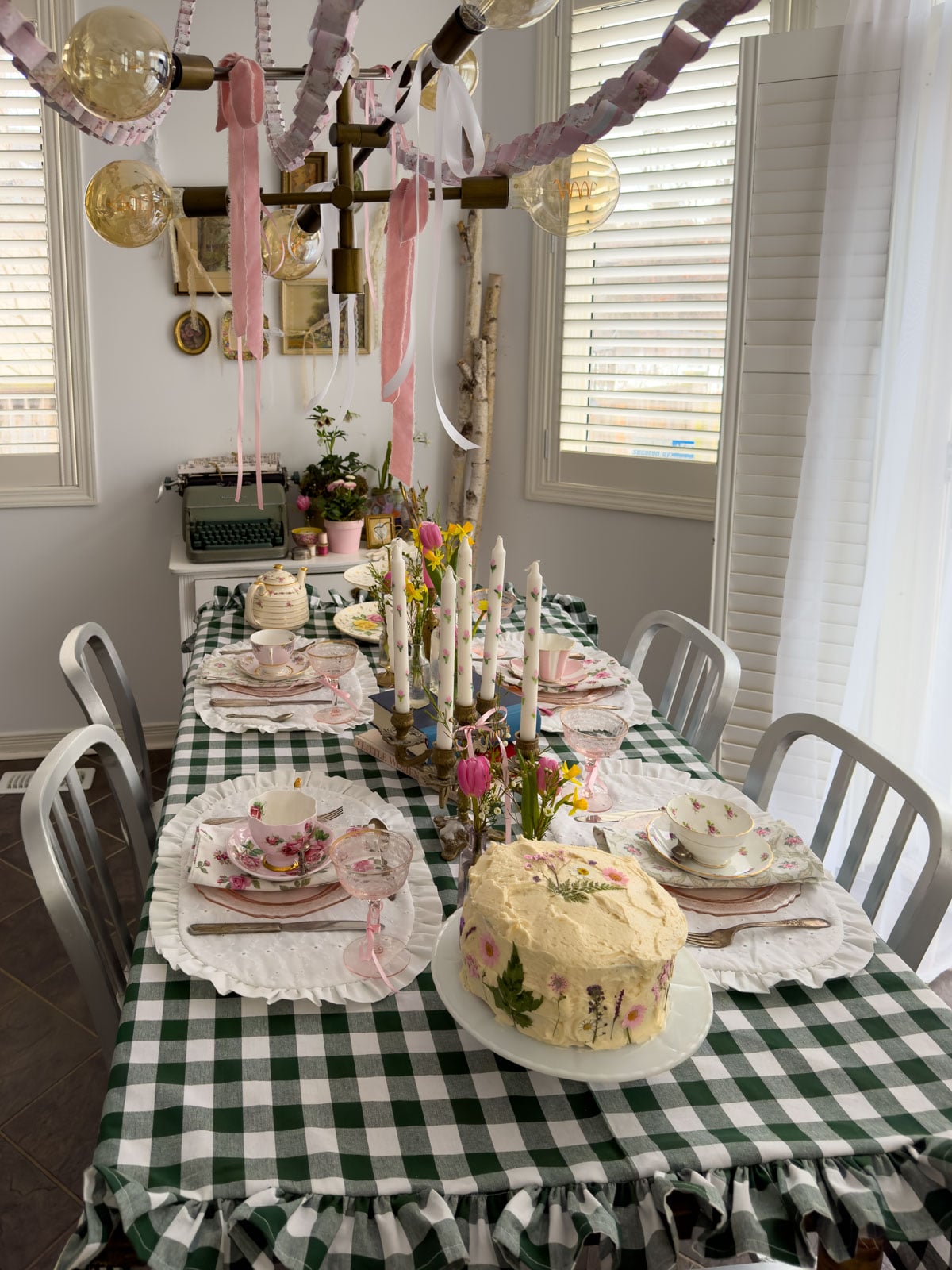 Long dining table set for a Little Women vintage tea party with paper chains overhead, hand-painted taper candles, pressed flower cake and vintage teacups on green gingham.