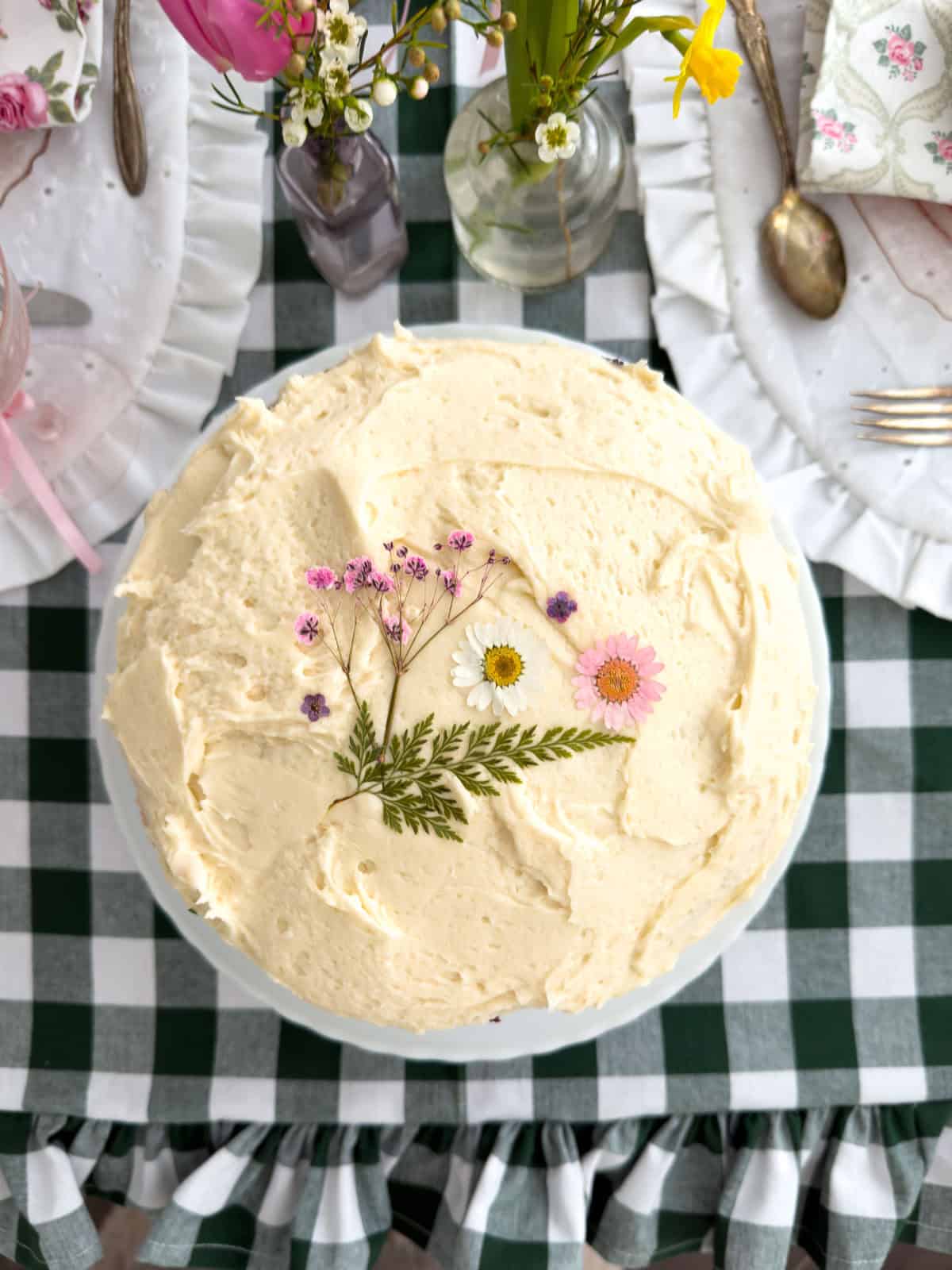 Overhead view of a Little Women party cake decorated with pressed daisies, pink flowers and a fern frond on a green gingham tablecloth.