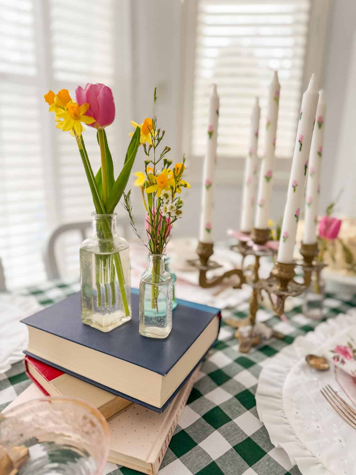 Glass bottles with pink tulips and daffodils on stacked books beside brass candelabra with hand-painted rose taper candles.