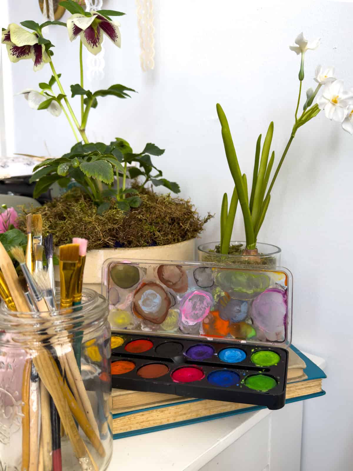 Mason jar of paintbrushes beside a used watercolor palette and spring flowers representing Amy March at a Little Women vintage tea party.