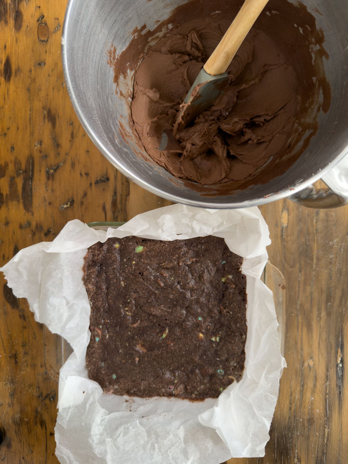 Baked mini egg brownies in a parchment lined pan beside a bowl of chocolate frosting ready to spread.