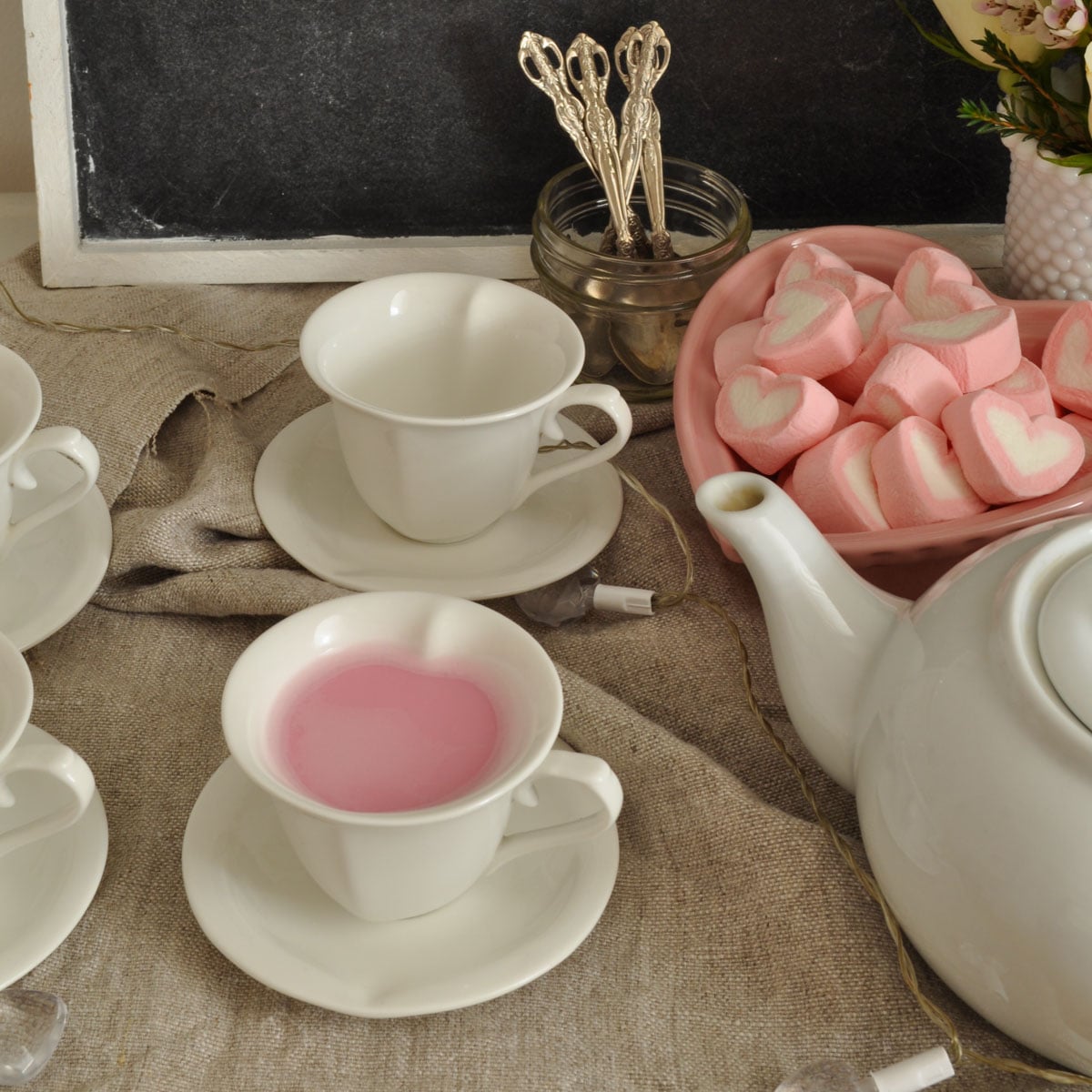 A white teacup filled with pink hot chocolate on a burlap runner beside a white teapot and a pink bowl of heart-shaped marshmallows.