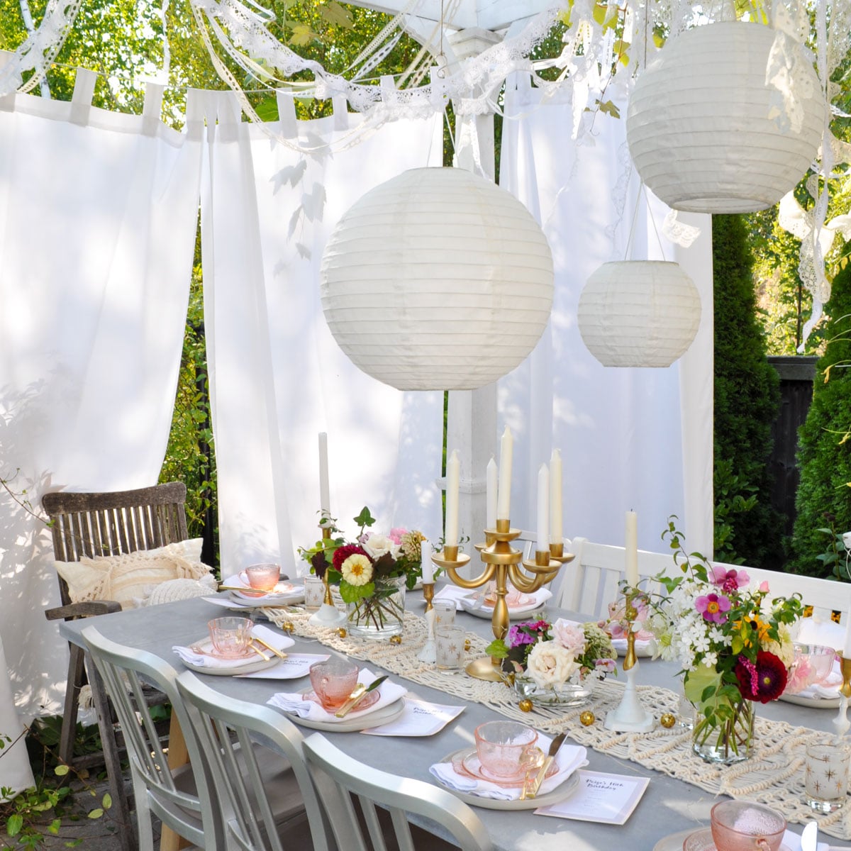 An outdoor summer tea party table set with pink glassware, gold candelabras, and fresh floral arrangements under a white draped pergola hung with oversized paper lanterns and lace garland.