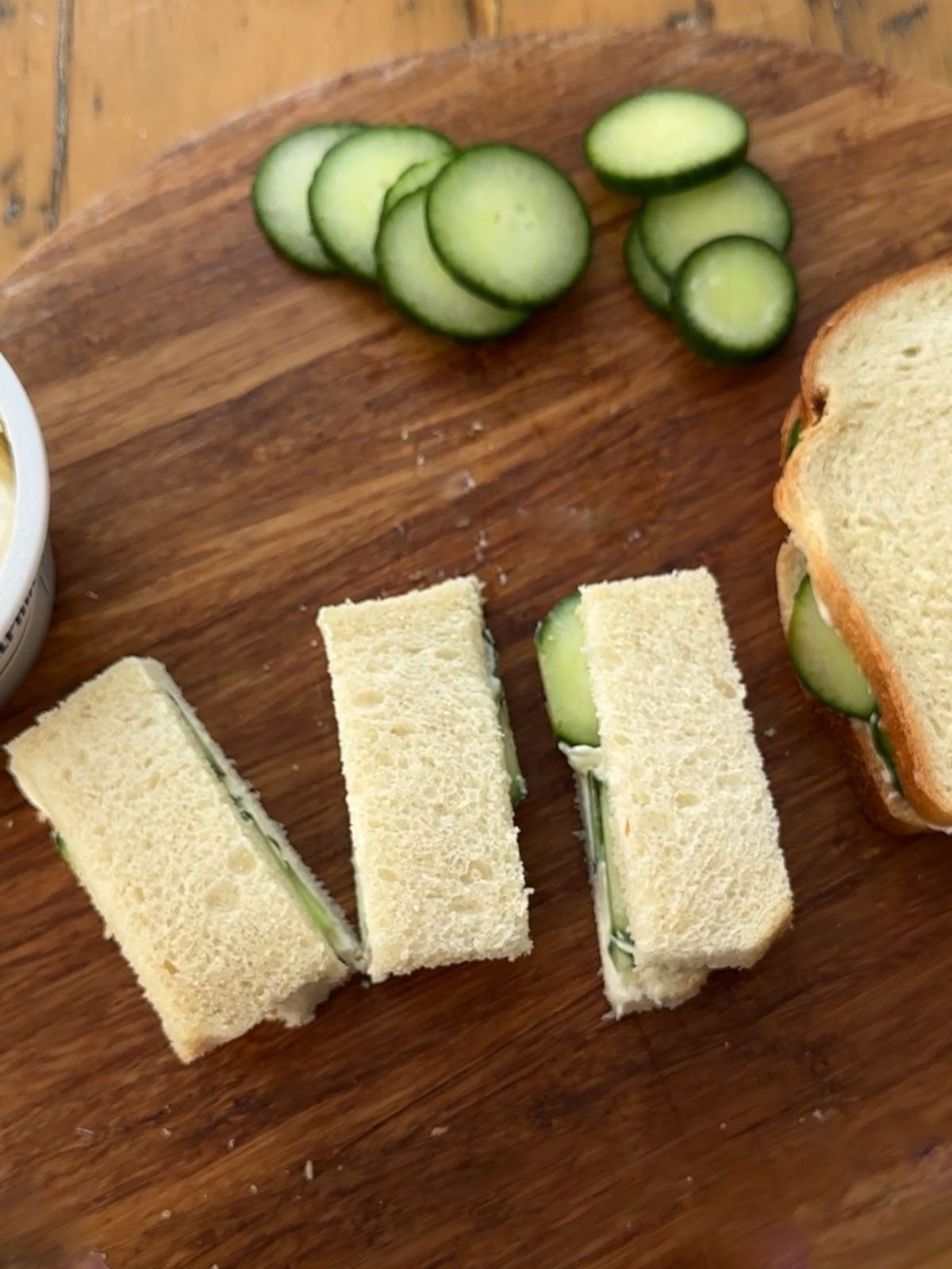 Three cucumber tea sandwich fingers cut and laid out on a round wooden cutting board beside the uncut fourth sandwich, with sliced English cucumber rounds and a container of cream cheese nearby.