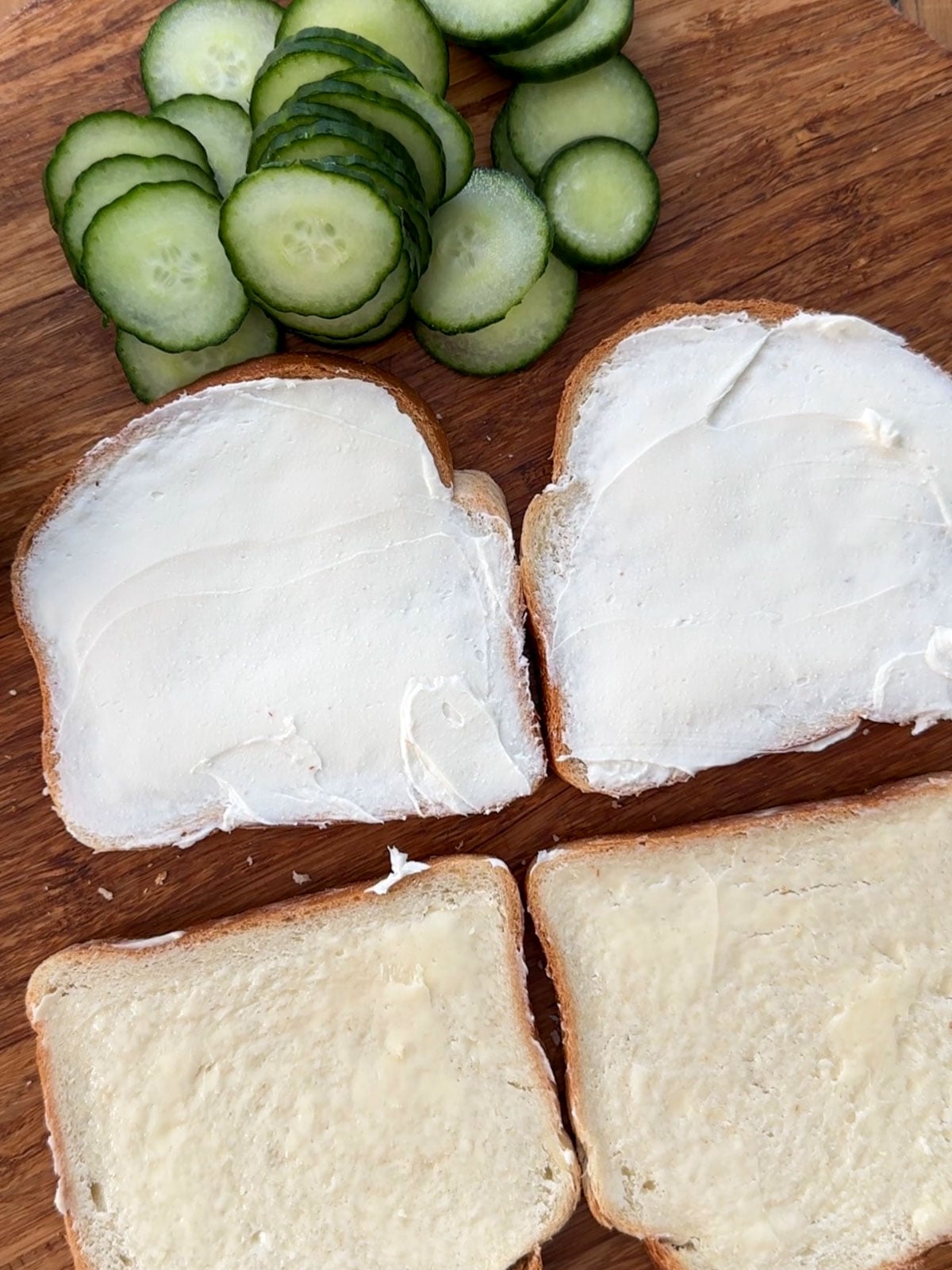 Two slices of white bread spread with cream cheese and two slices spread with butter on a round wooden cutting board with sliced cucumber rounds alongside.