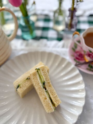 Two cucumber tea sandwich fingers on a white scalloped plate with a vintage floral teapot and a pink floral teacup of tea, pink tulips and bud vases on a green gingham tablecloth in the background.