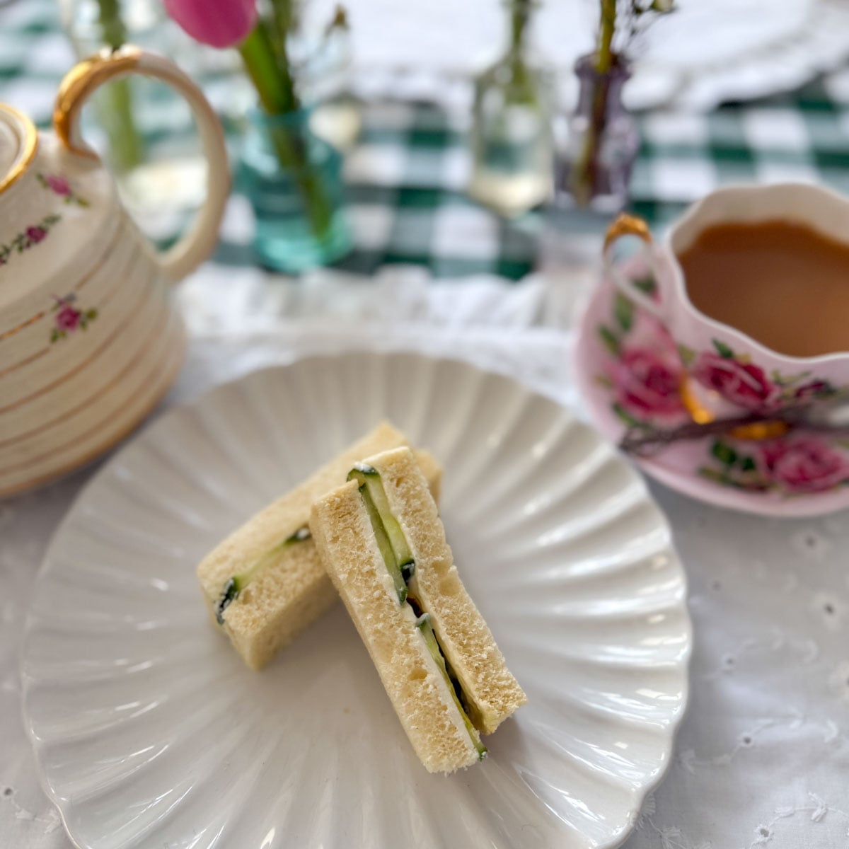 Two cucumber tea sandwich fingers on a white scalloped plate with a vintage floral teapot and a pink floral teacup of tea, pink tulips and bud vases on a green gingham tablecloth in the background.