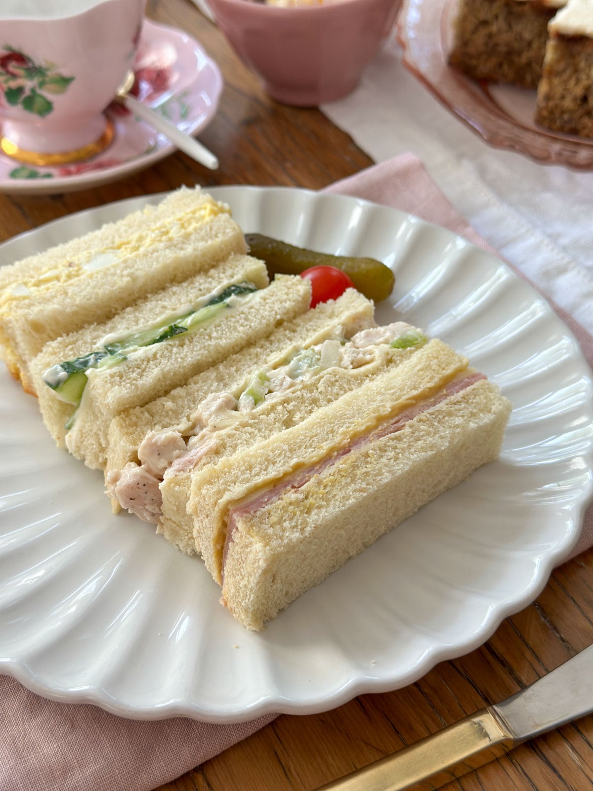 Tea party finger sandwiches including cucumber, egg salad, chicken salad, and ham on a white scalloped plate with a pink floral teacup and saucer in the background.