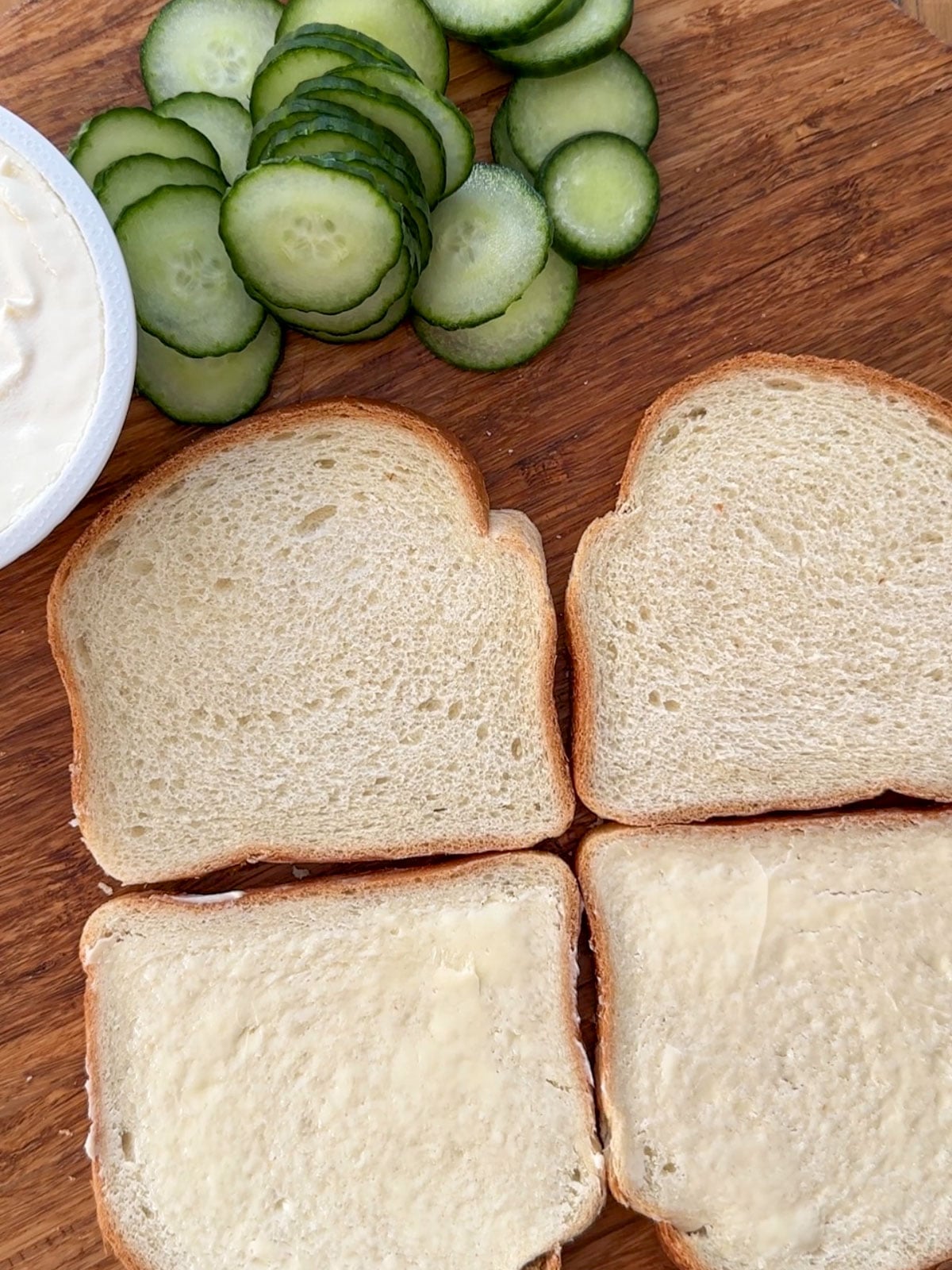 Four slices of white sandwich bread laid out on a round wooden cutting board with sliced English cucumber rounds alongside and a bowl of cream cheese.