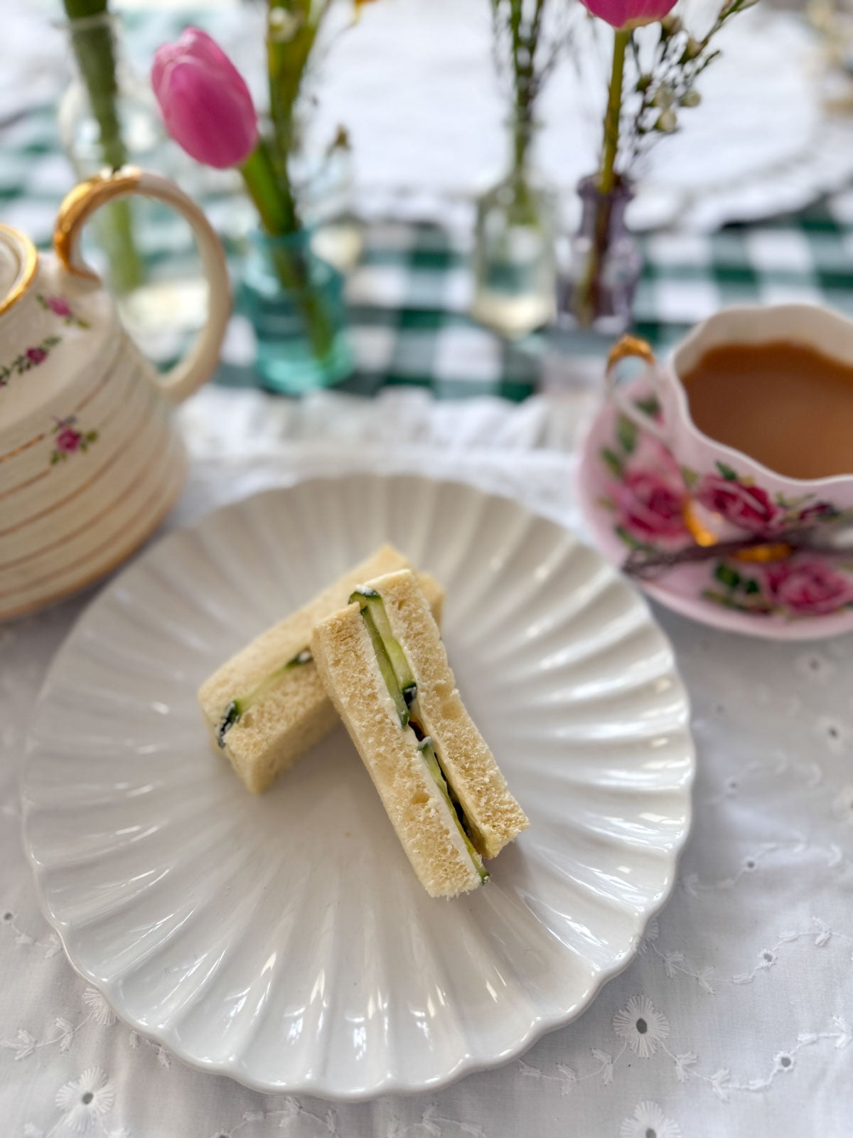 Two cucumber tea sandwich fingers on a white scalloped plate with a floral vintage teapot, a cup of tea with saucer, and pink tulips in the background.