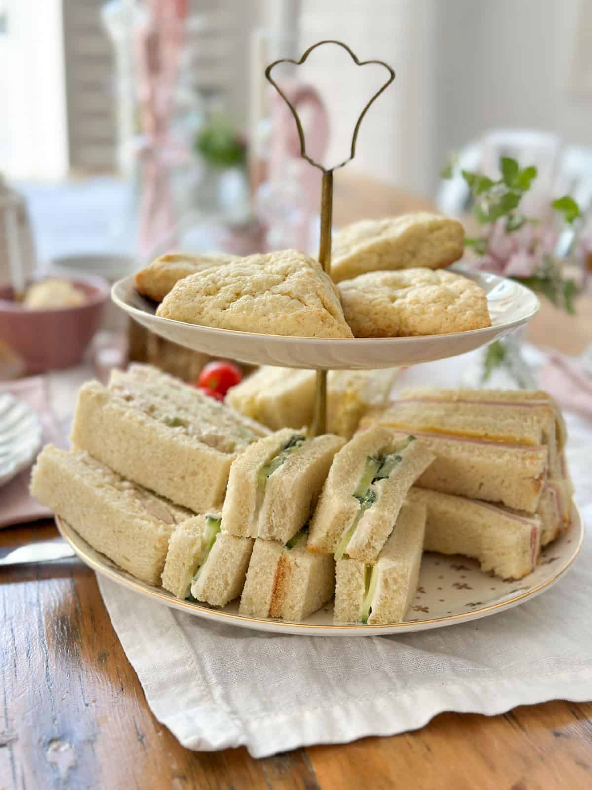Assorted tea party finger sandwiches and scones arranged on a two-tiered vintage china stand on a wooden table with a linen napkin.