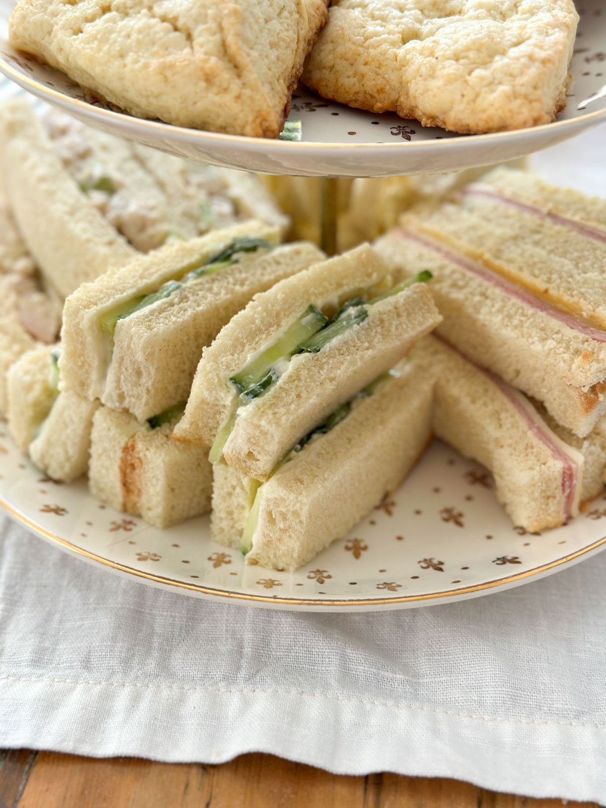 Cucumber tea sandwich fingers and ham tea sandwiches on the bottom tier of a vintage china two-tiered tray with scones on the upper tier.