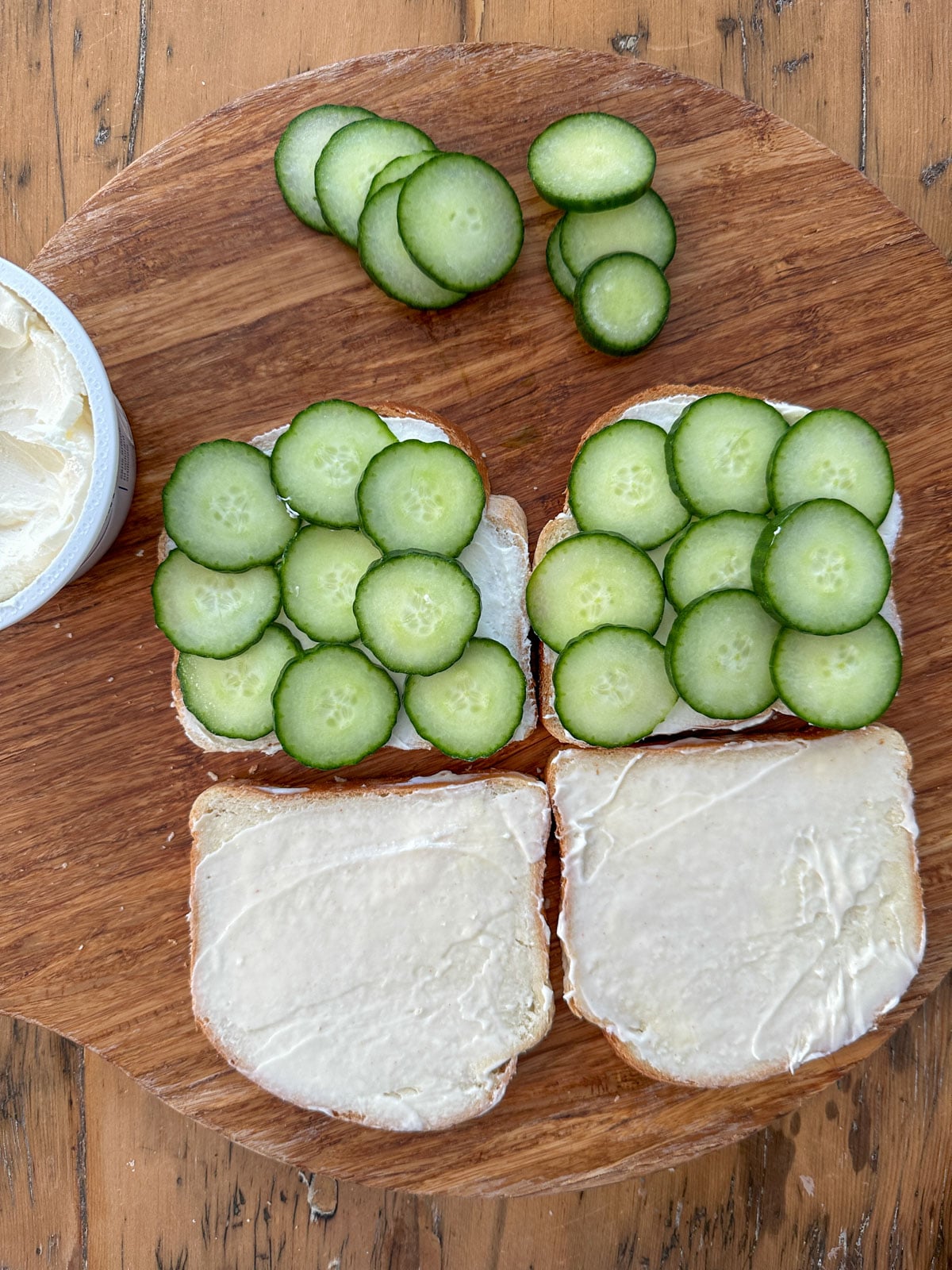 Thinly sliced English cucumber rounds layered over cream cheese spread on white bread slices on a round wooden cutting board.