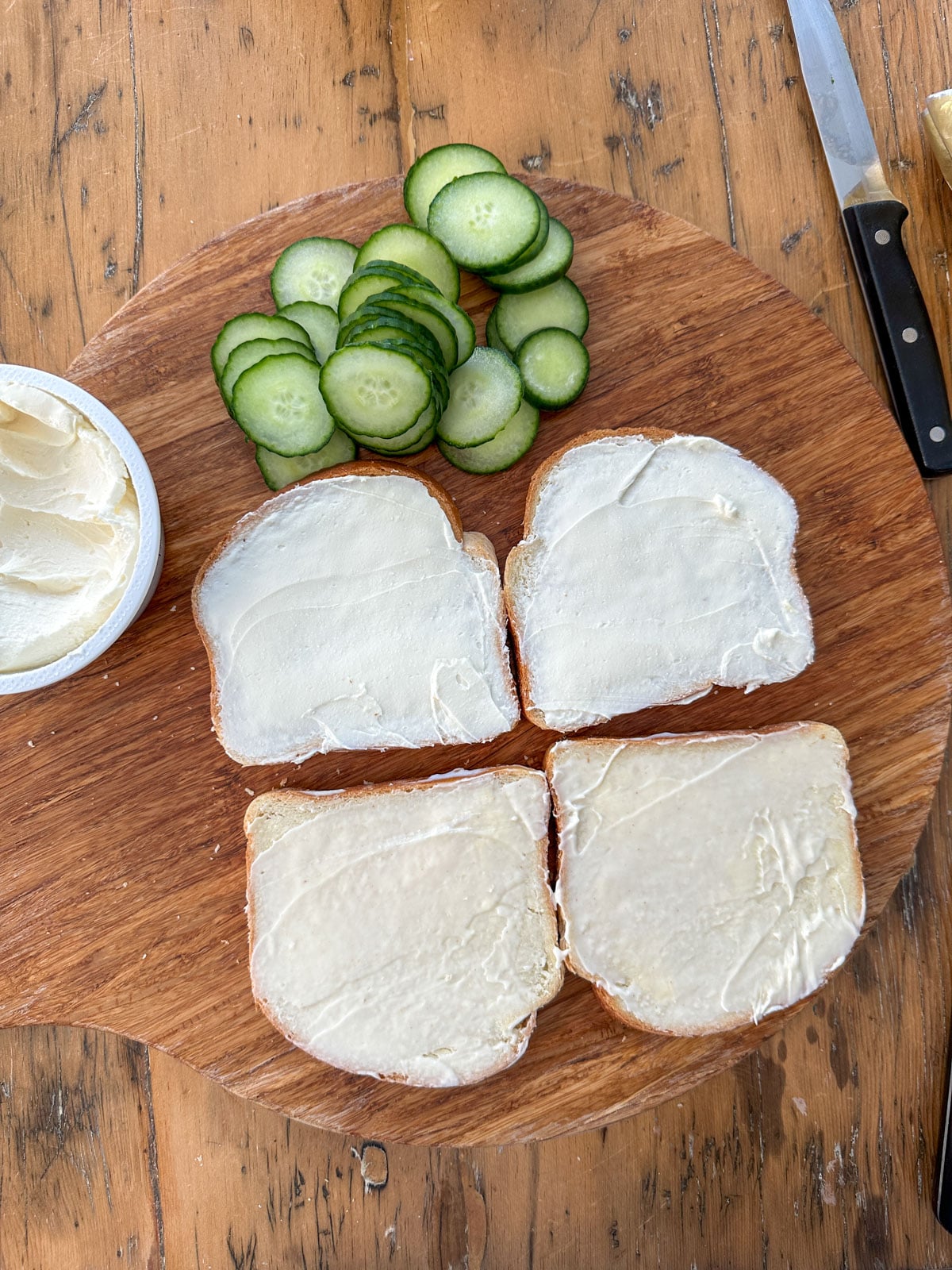 Four slices of white sandwich bread all spread with cream cheese or butter on a round wooden cutting board with sliced English cucumber rounds alongside.