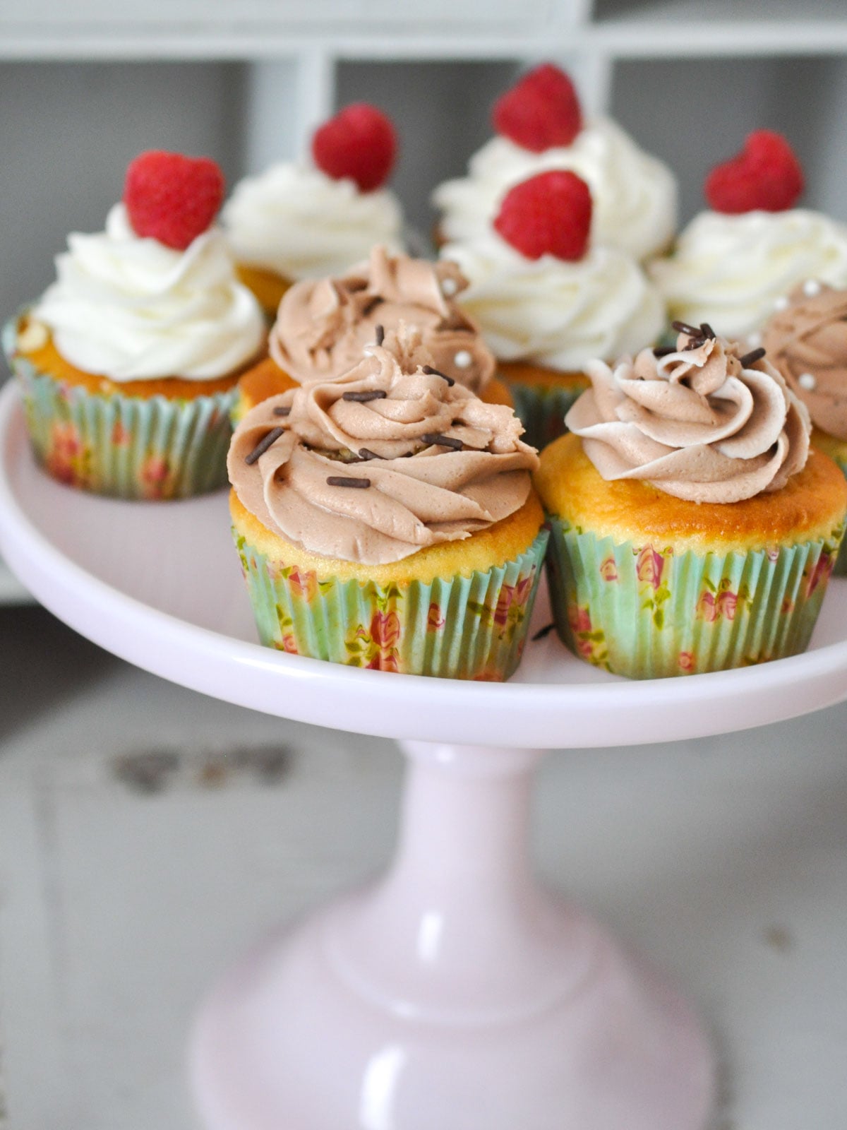 Cake plate of vanilla cupcakes, half with chocolate icing and sprinkles, and half with vanilla icing and raspberries.