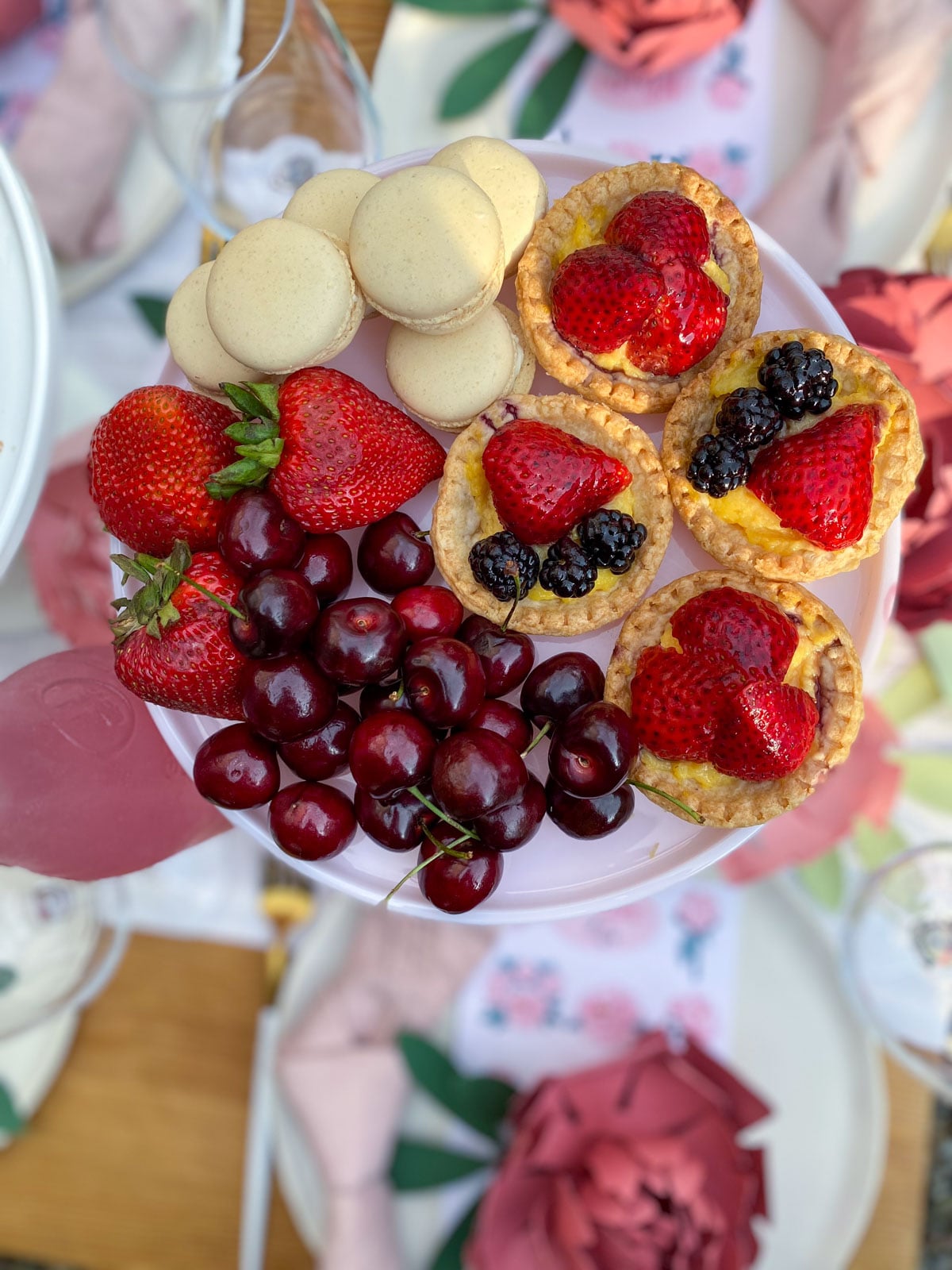 A pink cake stand topped with vanilla macarons, fresh fruit tarts filled with custard and topped with strawberries and blackberries, fresh cherries, and strawberries.