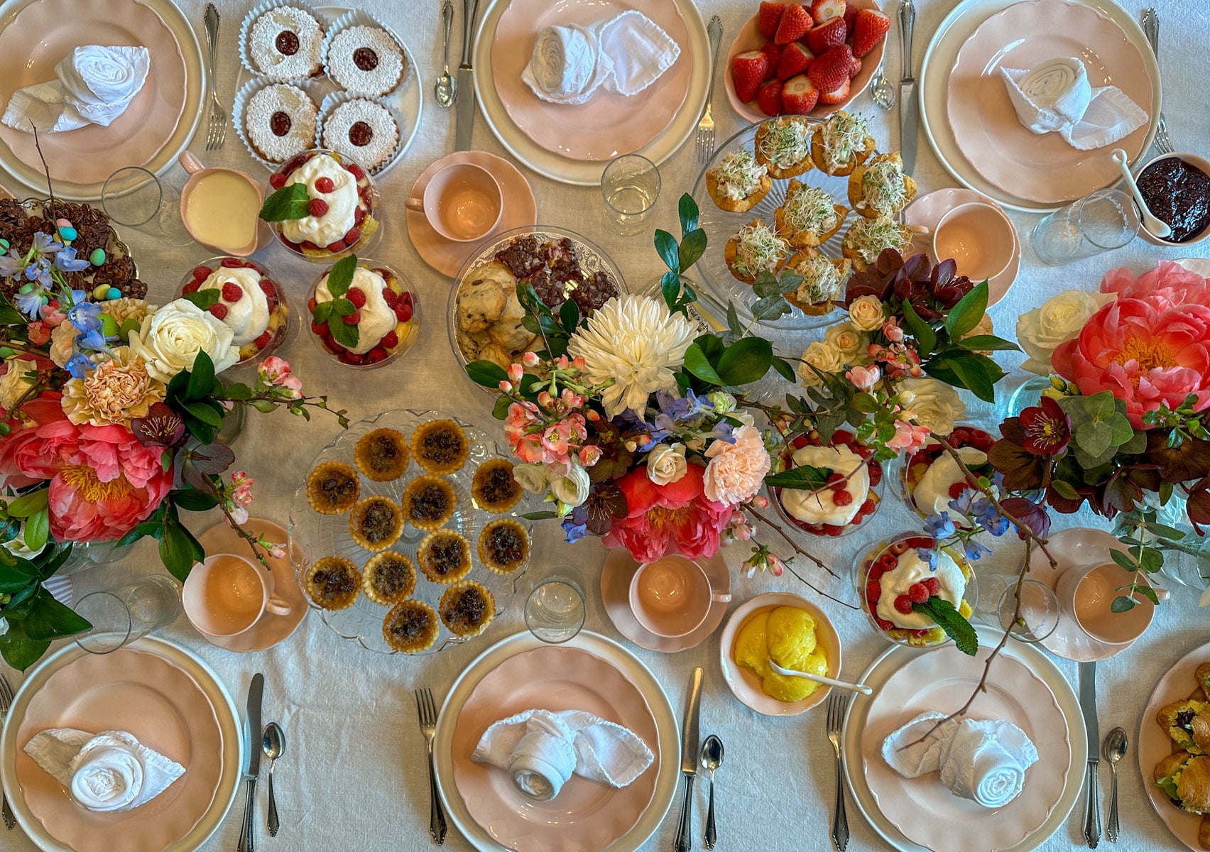 An overhead view of a tea party table set with pink dishes and linen napkins, loaded with butter tarts, scones, raspberry trifles, jam cookies, and chicken salad sandwiches surrounded by lush floral centerpieces of peonies, roses, and dahlias.