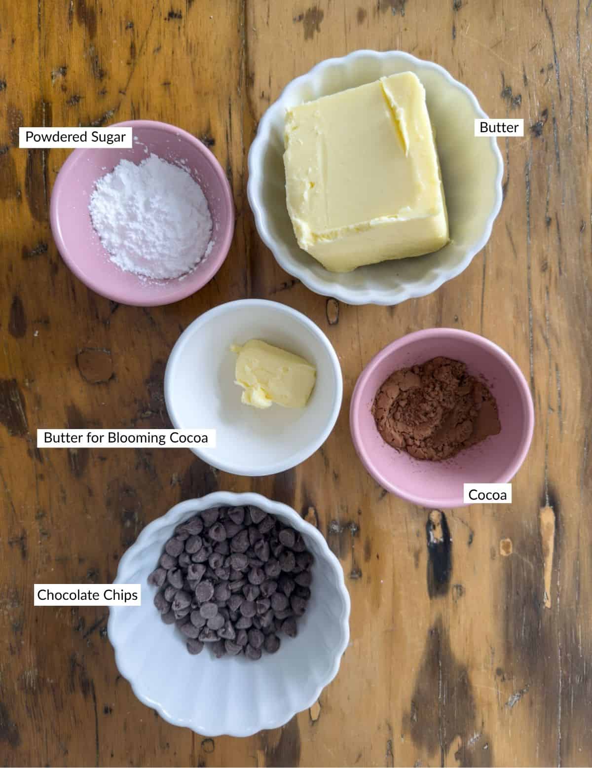 Ingredients for chocolate butter including butter, chocolate chips, cocoa powder, powdered sugar, and a small pat of butter arranged in small bowls on a wooden table.