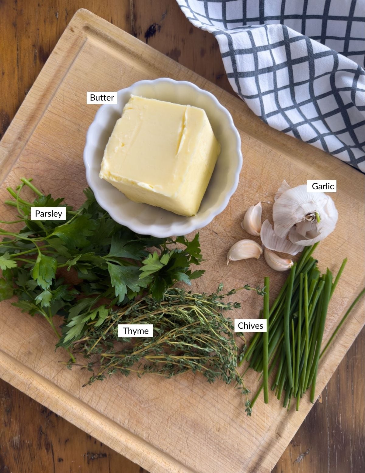Ingredients for garlic herb compound butter including a block of salted butter, fresh flat leaf parsley, thyme, chives, and garlic on a wooden cutting board.