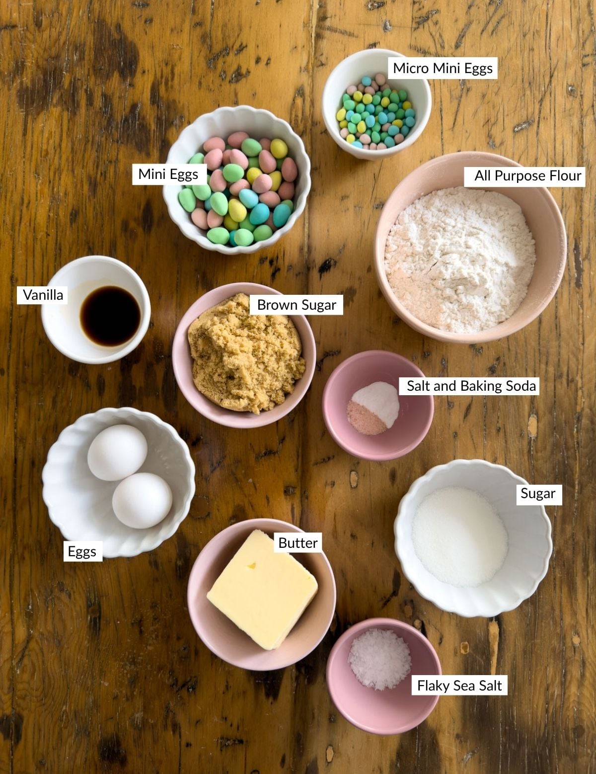 Ingredients for Cadbury mini egg cookies including butter, brown sugar, flour, eggs, vanilla, and both regular and micro Cadbury mini eggs arranged in bowls on a wood table.