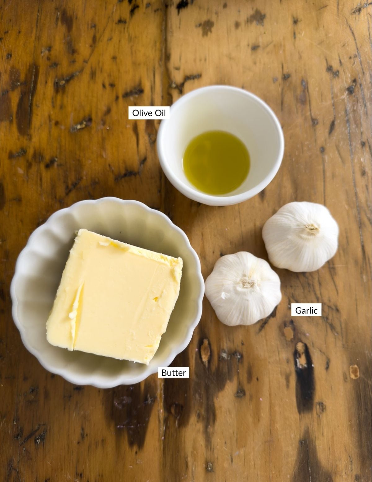Overhead view of a block of butter on a scalloped plate, olive oil in a white bowl, and two heads of garlic on a wooden table.