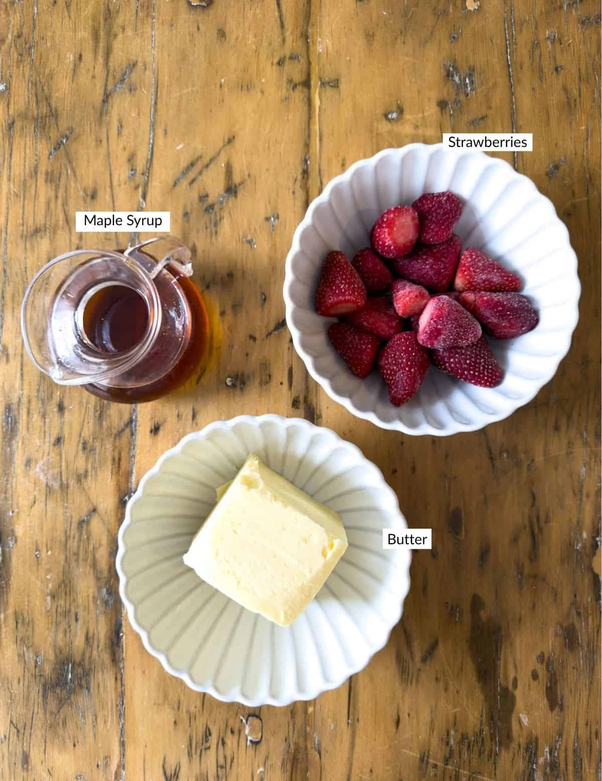 Overhead view of strawberry butter ingredients on a wood table: a glass pitcher of maple syrup, a white scalloped bowl of frozen strawberries, and a white scalloped bowl with a block of butter, each labeled.