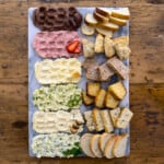 Overhead view of a compound butter board with six flavored compound butters in a row including chocolate, strawberry, whipped honey, jalapeño honey, roasted garlic, and garlic herb, each paired with its own bread on a marble board.