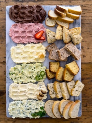Overhead view of a compound butter board with six flavored compound butters in a row including chocolate, strawberry, whipped honey, jalape&ntilde;o honey, roasted garlic, and garlic herb, each paired with its own bread on a marble board.