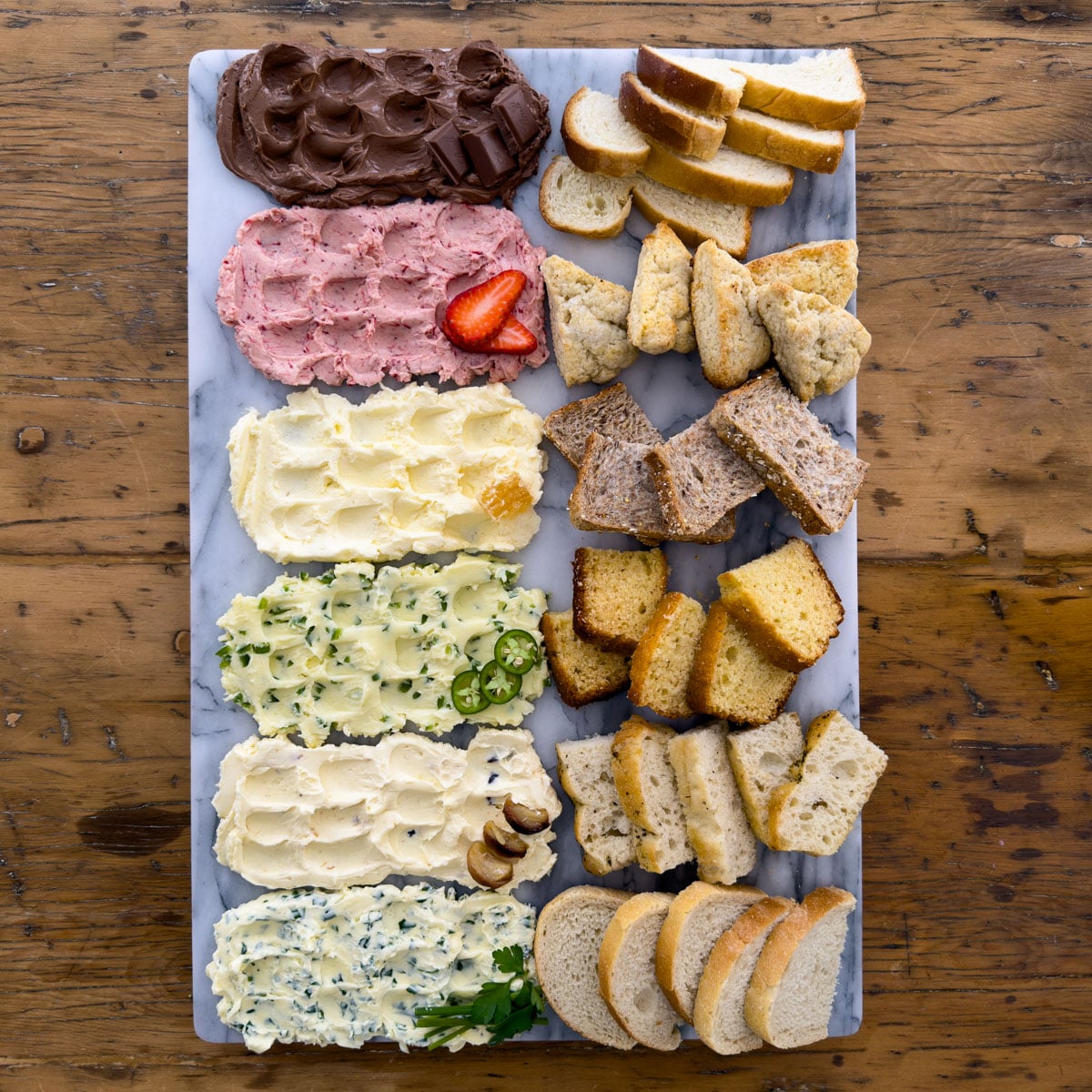 Overhead view of a compound butter board with six flavored compound butters in a row including chocolate, strawberry, whipped honey, jalape&ntilde;o honey, roasted garlic, and garlic herb, each paired with its own bread on a marble board.