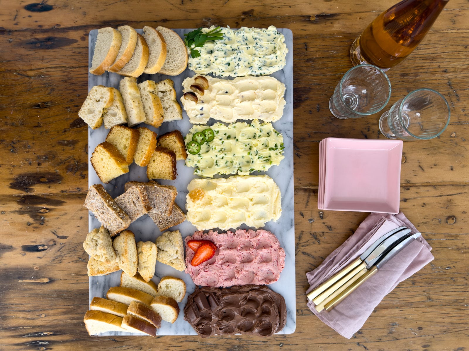 Overhead view of a complete compound butter board with six compound butters and paired breads on a marble board, styled with pink plates, gold butter knives, linen napkins, and wine glasses on a wood table