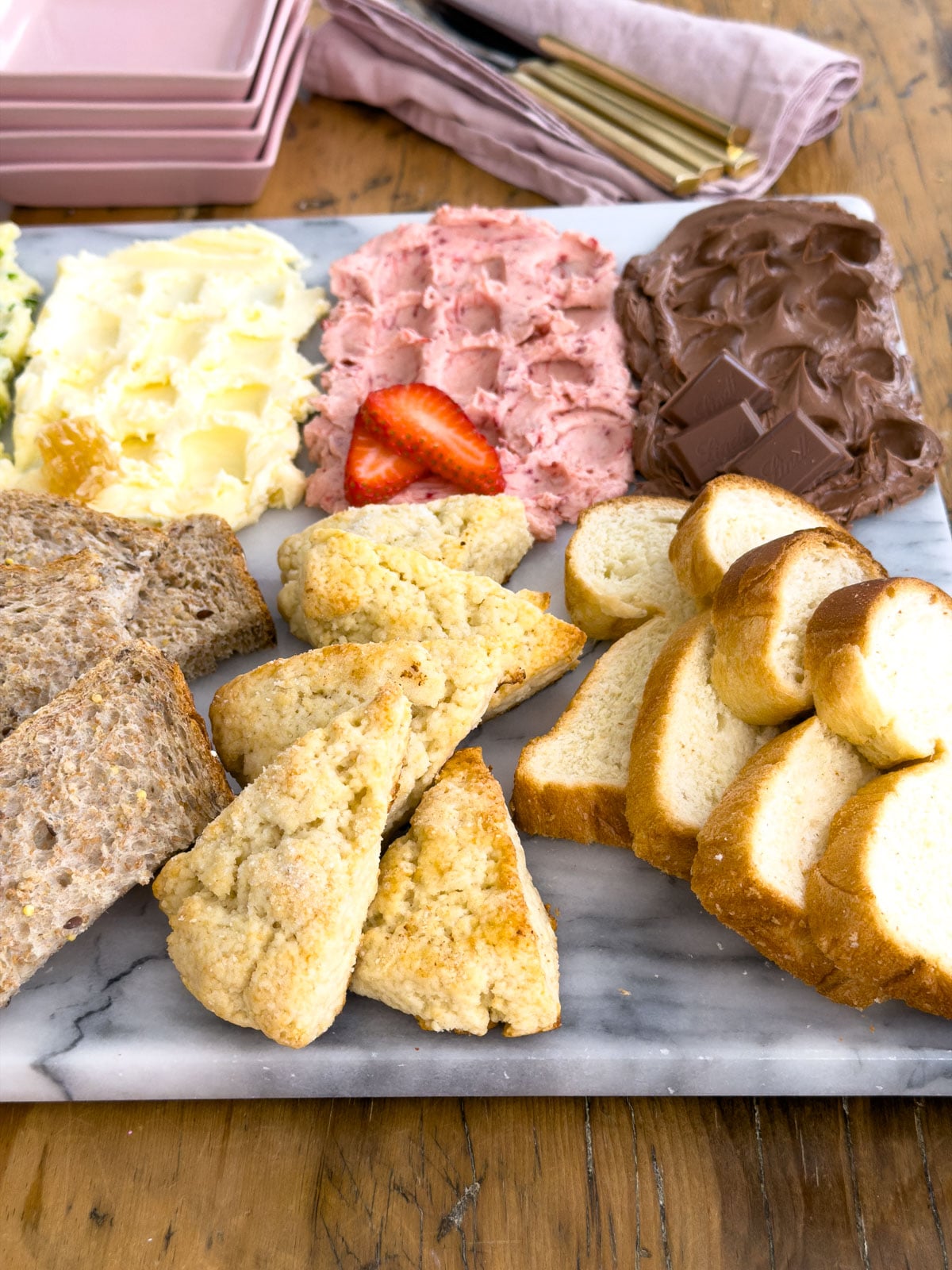 Scones and sliced brioche arranged on a marble butter board alongside strawberry compound butter and chocolate compound butter, showing the bread pairings for the sweet section of the butter flight