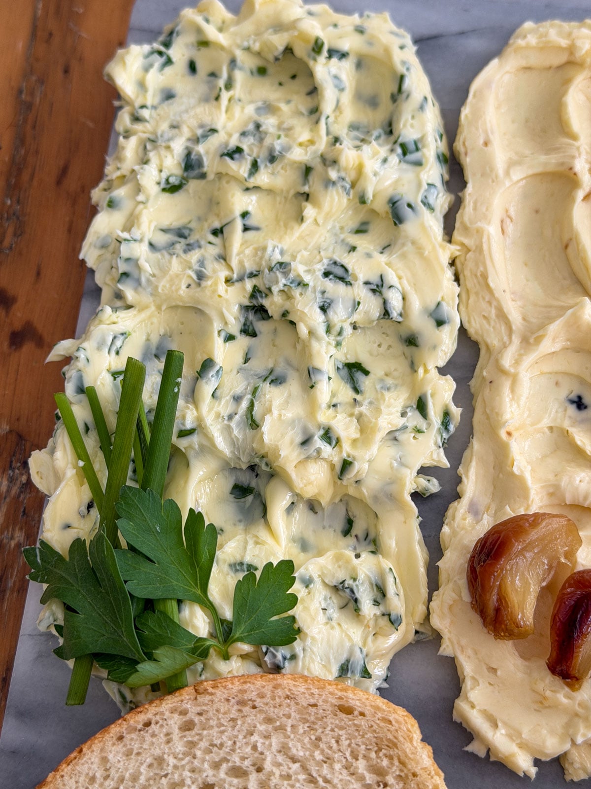 Close up of garlic herb compound butter spread on a marble board with fresh chives and flat leaf parsley garnish and sliced sourdough bread alongside.