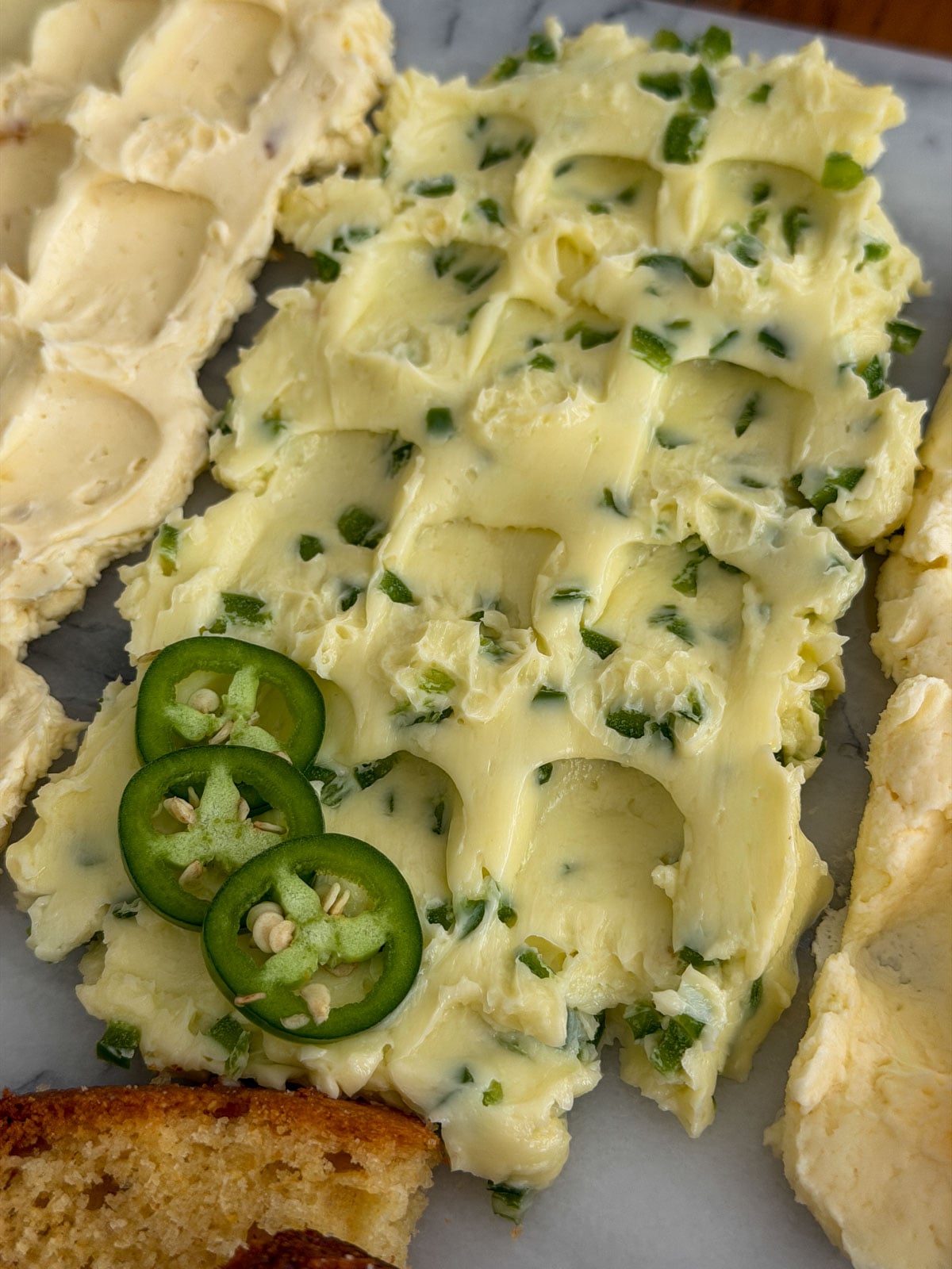 Close up of jalape&ntilde;o honey compound butter spread on a marble board with fresh jalape&ntilde;o rings as garnish and cornbread slices alongside.