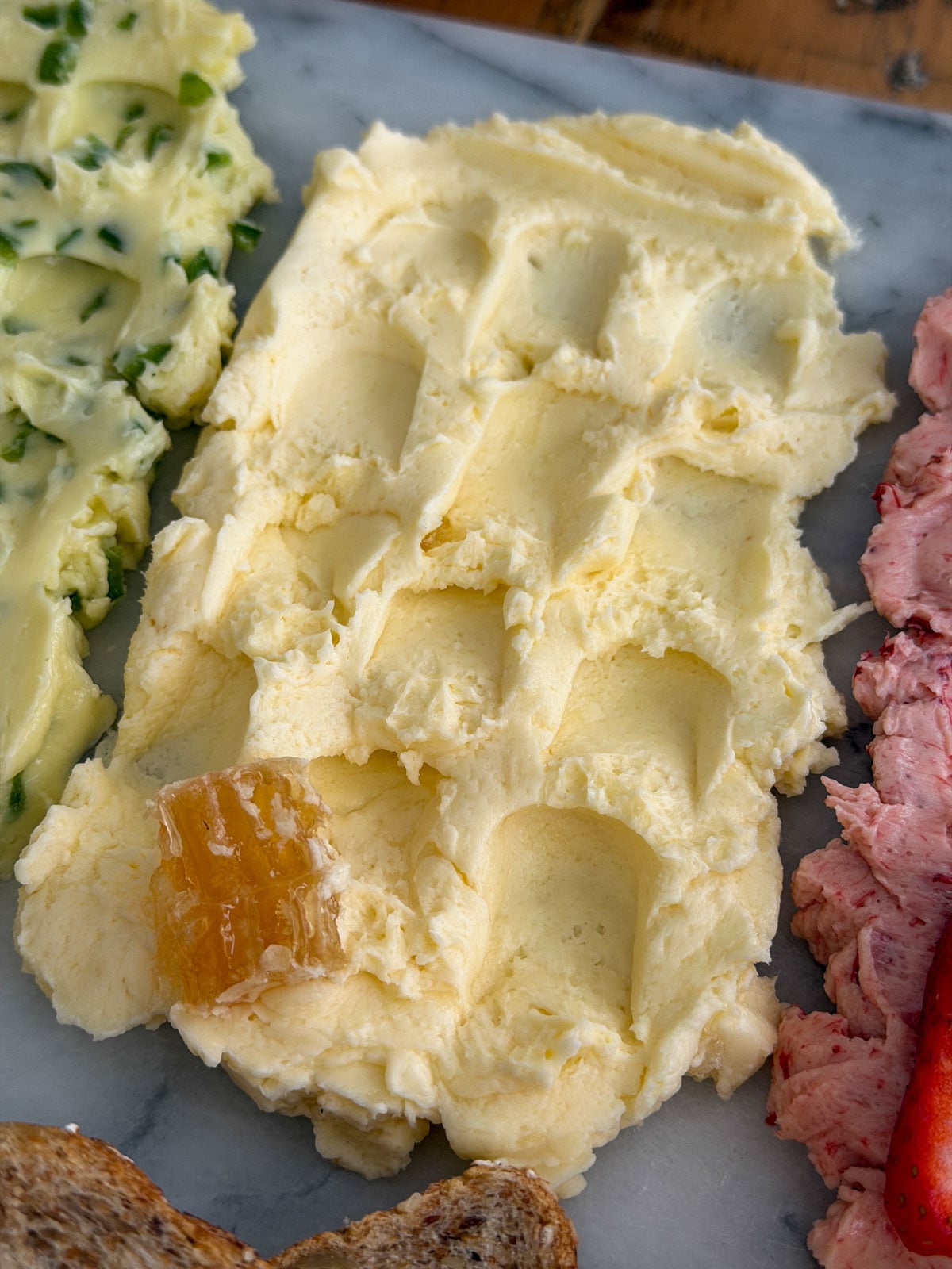 Close up of whipped honey compound butter spread on a marble board with a piece of golden honeycomb as garnish and seeded oat bread alongside